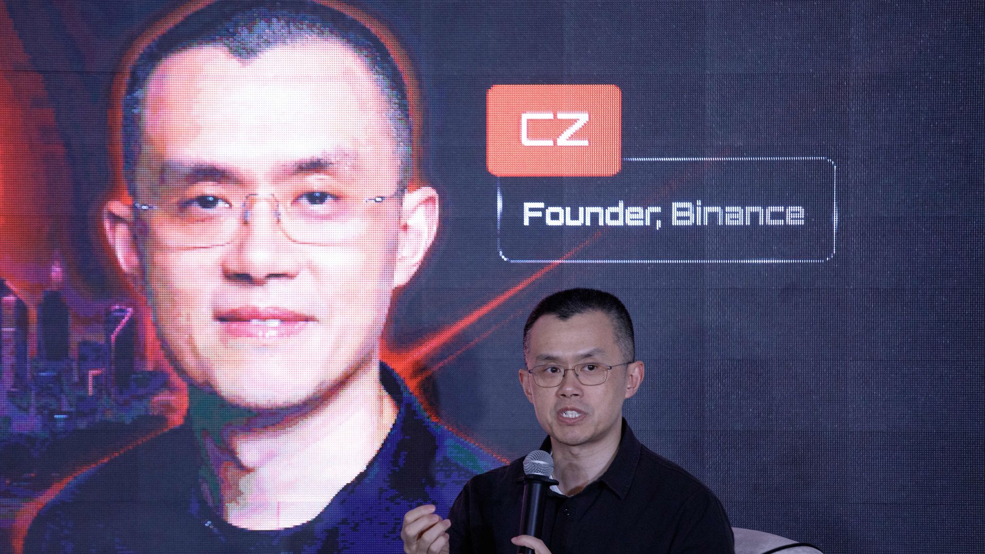 A man in a black shirt and glasses, holding a microphone, seated in front of a larger picture of himself. 