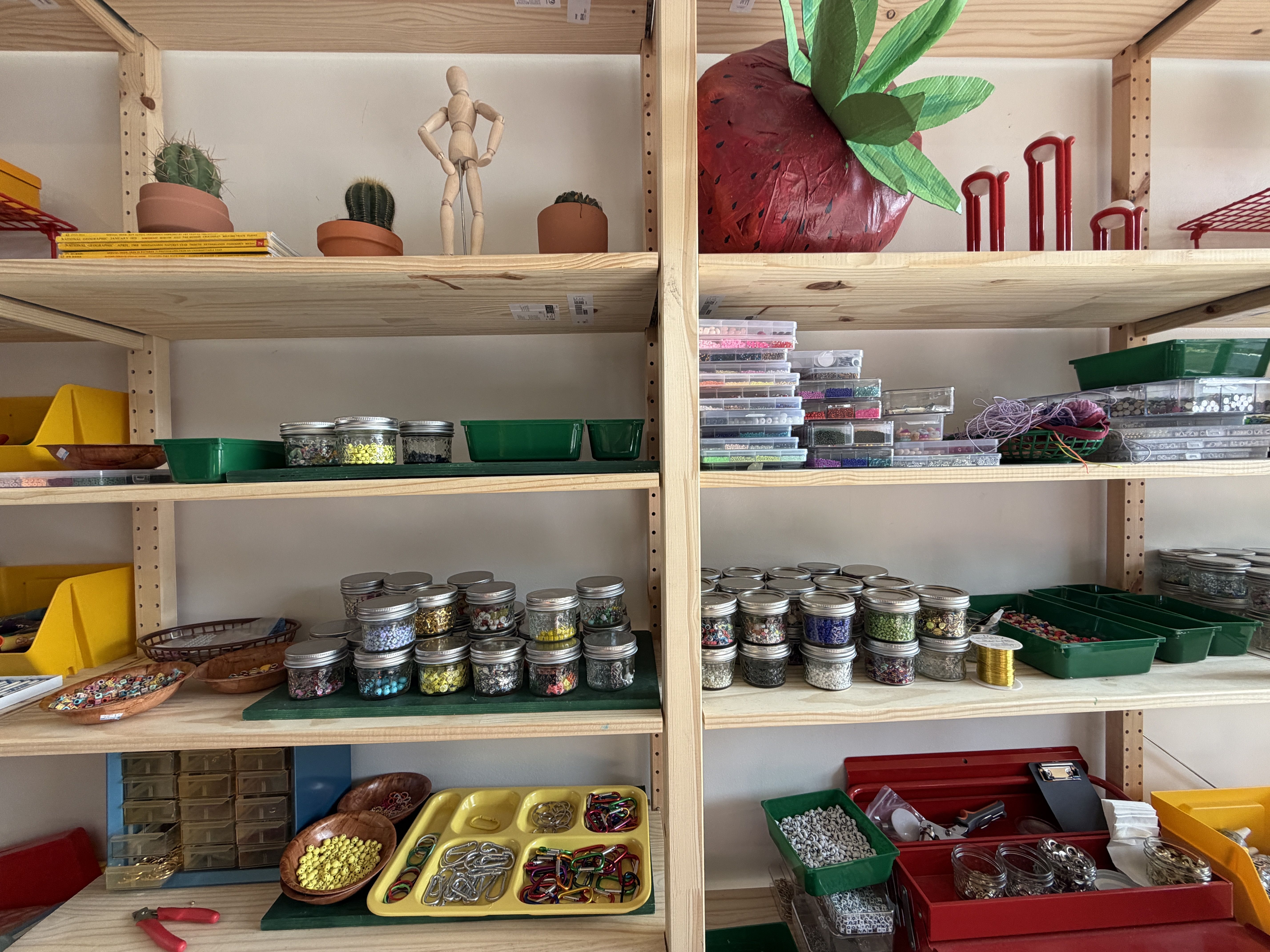 Wooden shelves filled with jars of beads, small colored items, green trays, a wooden mannequin, three small cactus plants, and a large decorative red strawberry with green leaves.