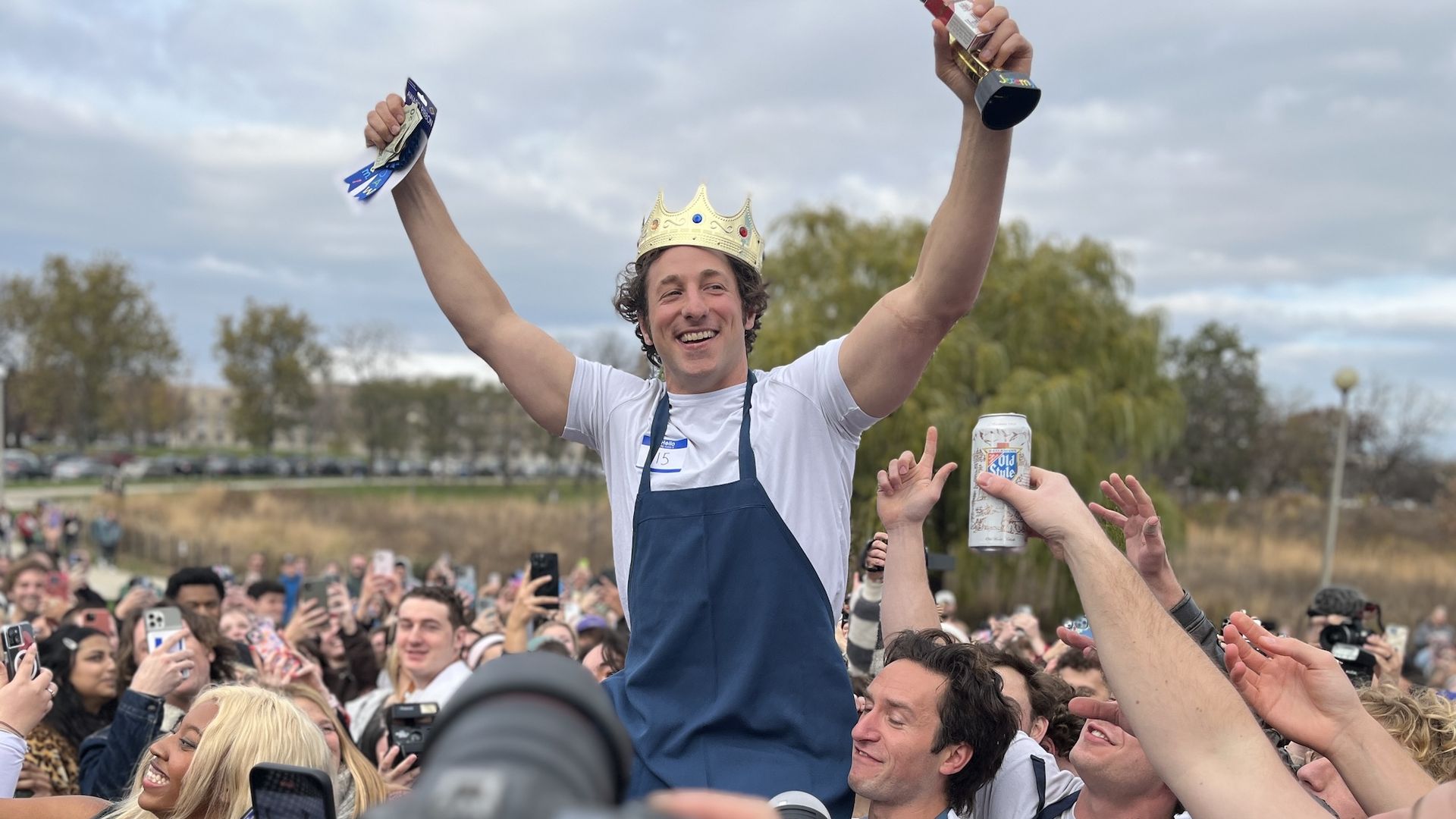 A man in a white t-shirt and blue apron is hoisted on a crowd's shoulders holding money in one hand and a trophy in another wearing a gold crown.