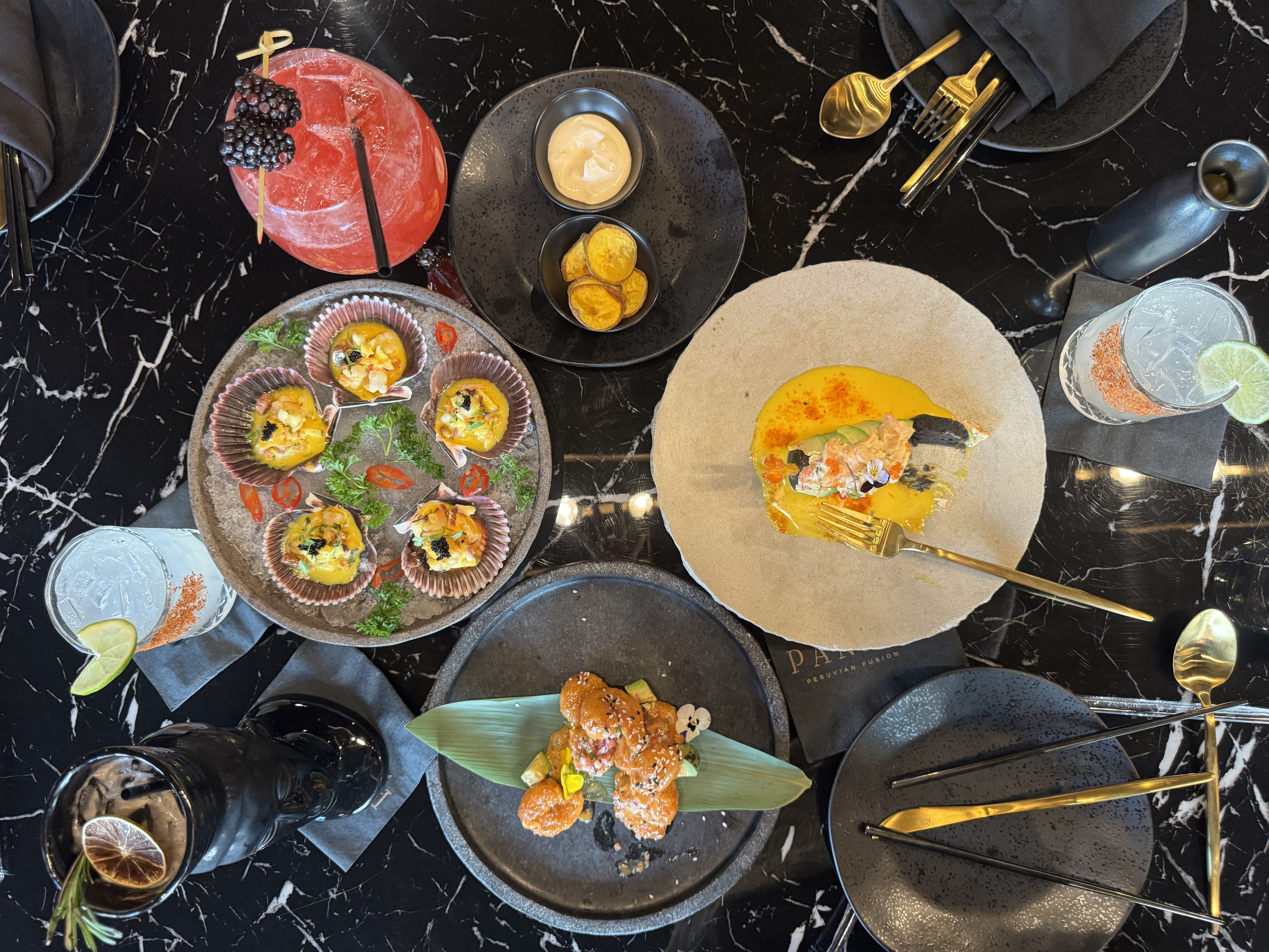 Overhead view of a black marble table with gourmet dishes, including scallops in shells with yellow sauce, a dish with yellow sauce and a gold fork, and a plate with fried bites on a green leaf. Drinks with lime and blackberries and gold and black utensils are also on the table.
