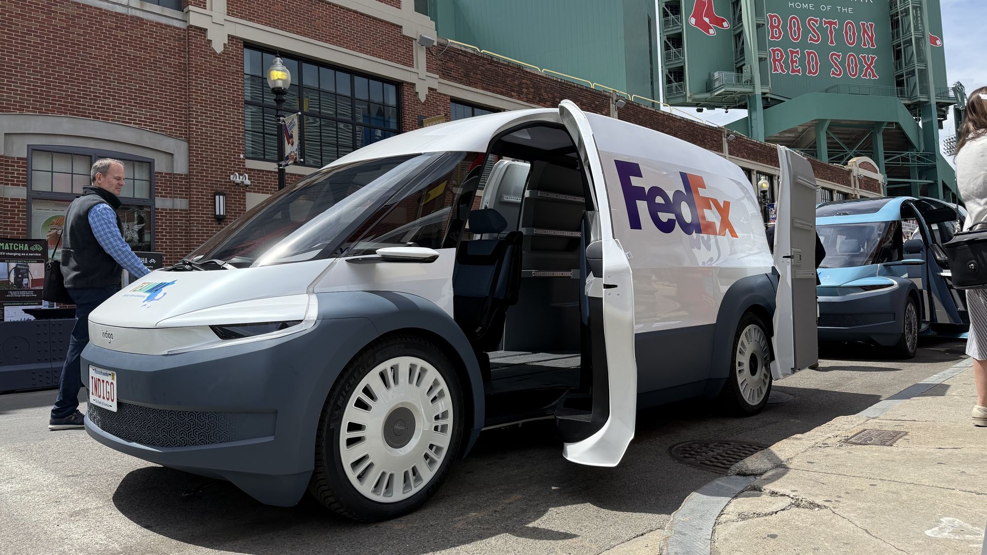 A photo of two Indigo Technologies electric delivery vans, taken from the front driver's-side quarter panel, parked on the street outside Fenway Park.