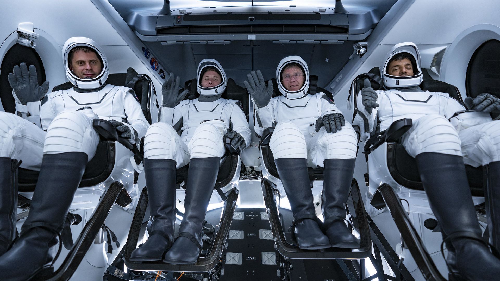 Four crewmembers sitting in a SpaceX Crew Dragon in white suits waving at the camera