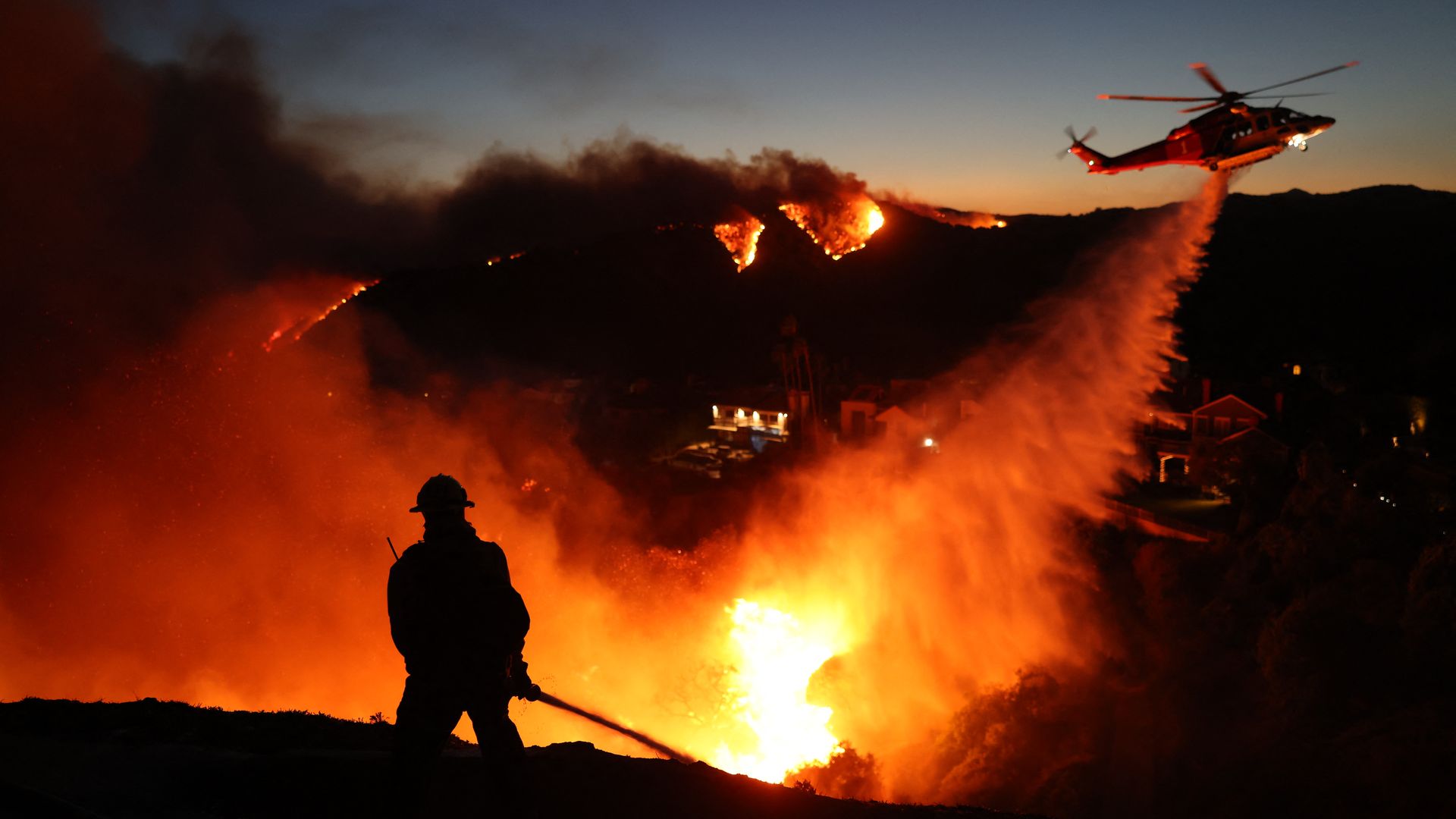 A firefighter battles a blaze as a helicopter hovers nearby.