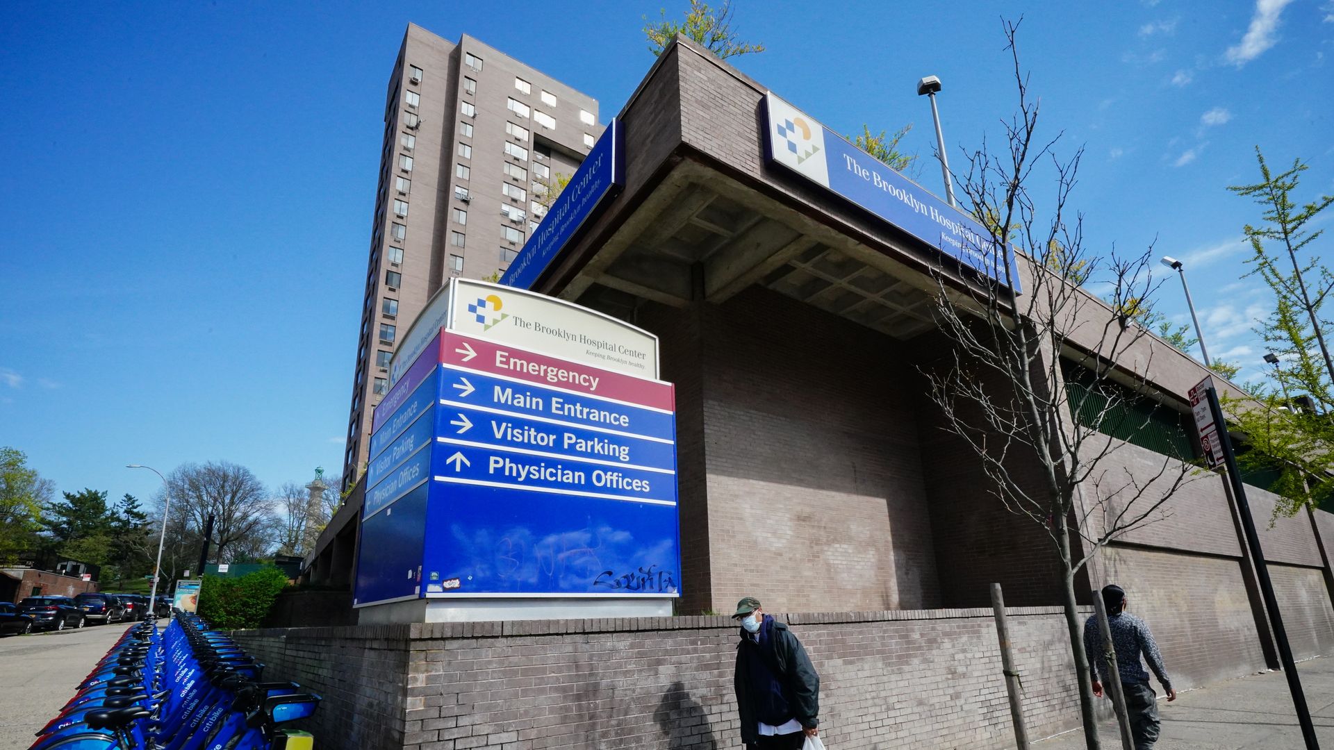A blue Brooklyn Hospital Center sign with people walking in front and the hospital in the background.