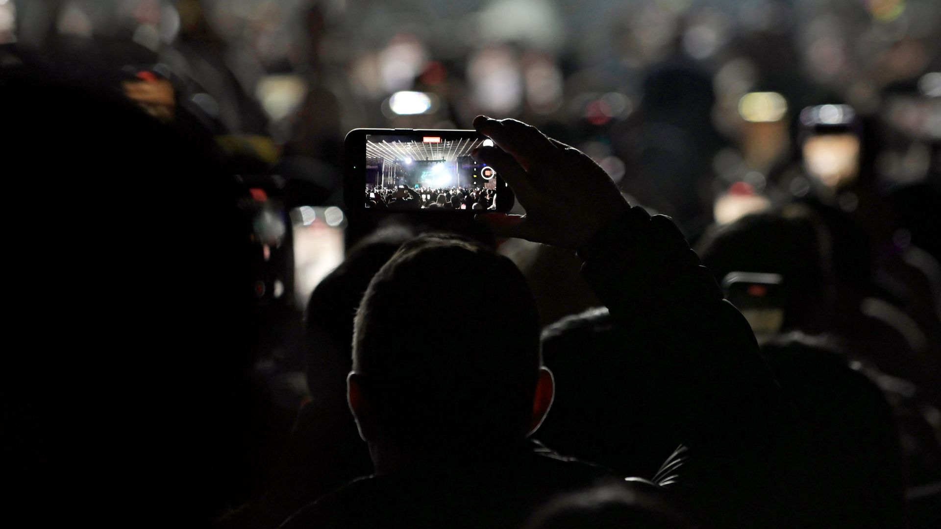 A person in a dark crowd holds up a smartphone to record a concert. The phone screen shows a bright stage with performers and lights, surrounded by blurred silhouettes of other concertgoers.