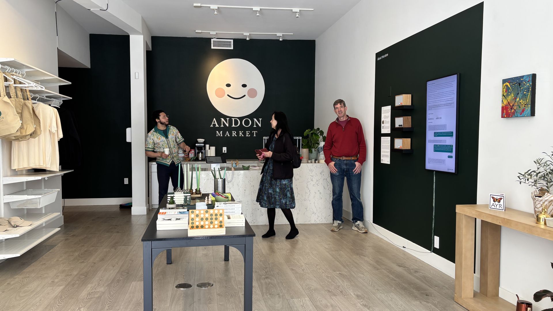 Interior of Andon Market: dark green wall with a large smiling circle logo, three people by a marble counter, left shelves with beige clothing, a center table, and a right digital display.