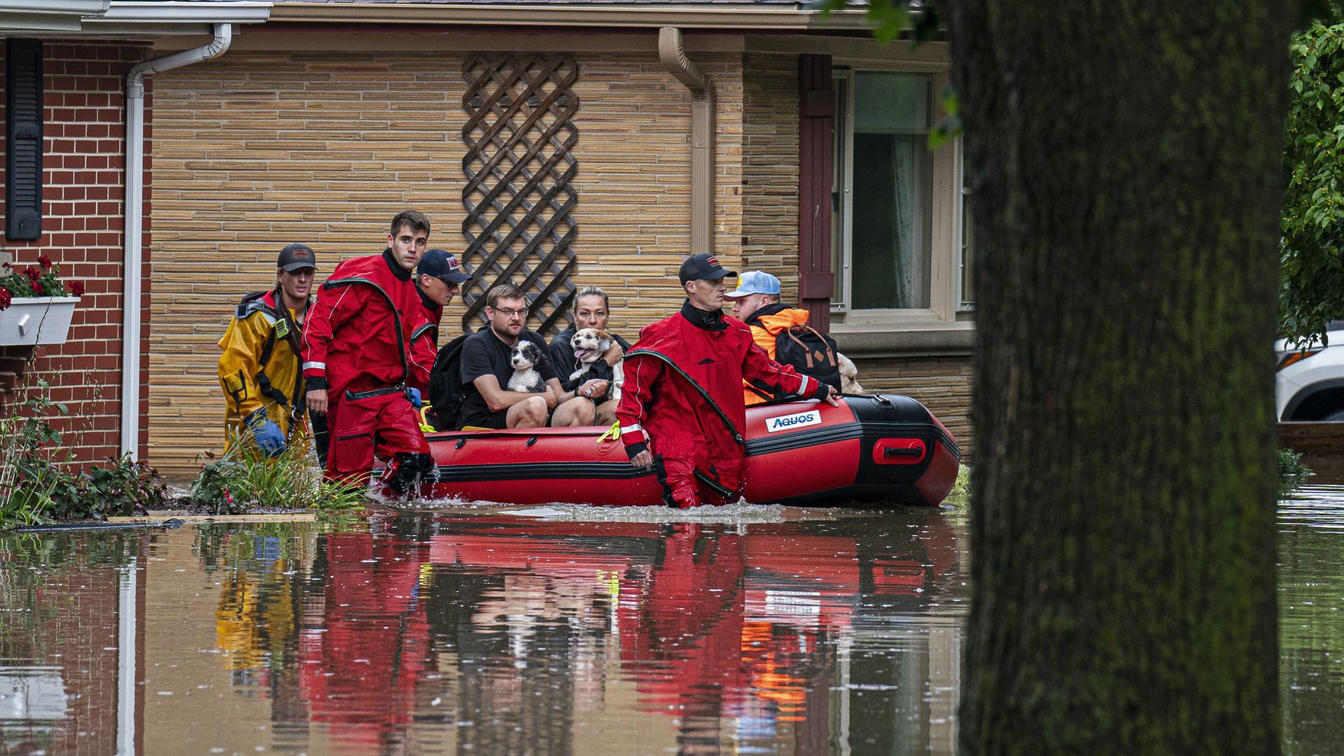 Rescue workers in red and yellow suits evacuate people and dogs by red inflatable boat through floodwater in a suburban area with brick and tan houses.