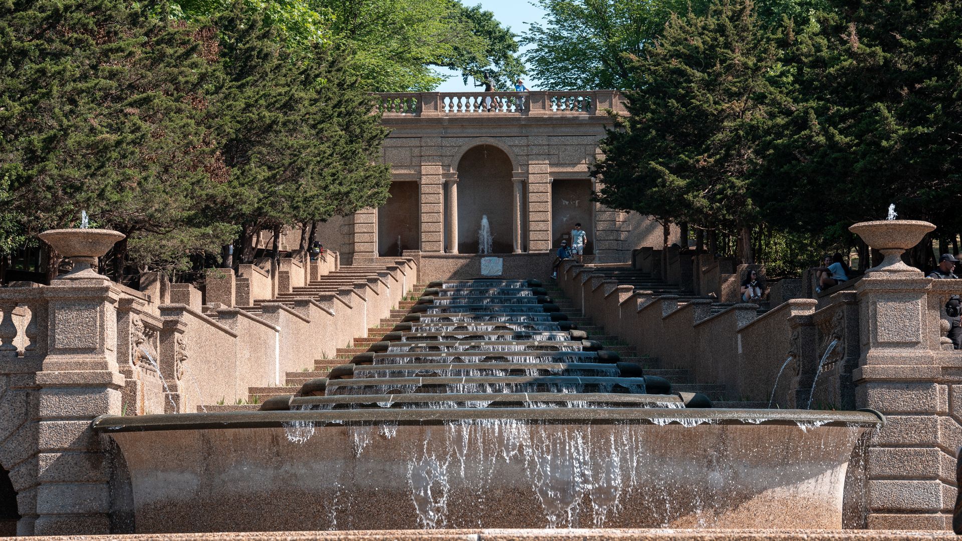 Symmetrical stone staircase fountain framed by trees, leading to an arch pavilion with a balcony. Water cascades down the steps as visitors sit along the sides and a few stand on the top level.