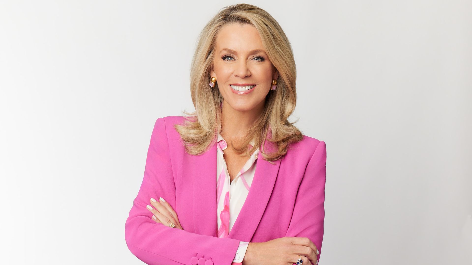 Smiling woman with blonde hair wearing a bright pink blazer and patterned blouse, arms crossed, against a plain light gray background.