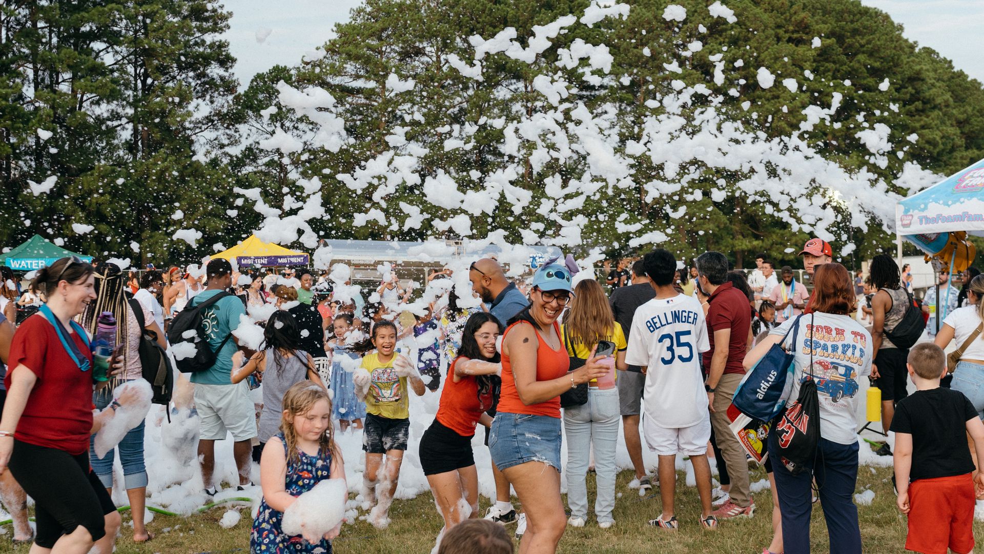 Crowd of diverse people, adults and children, playing in foam at outdoor event on grass with trees and tents in background under light blue sky.