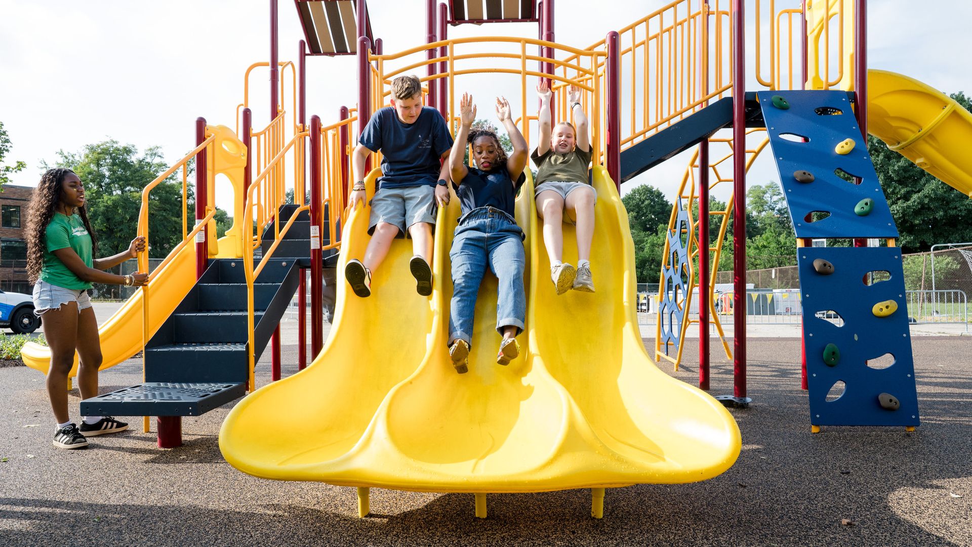 Kids slide down a bright yellow playground slide, surrounded by orange rails and a blue climbing wall; a girl in a green shirt watches nearby.