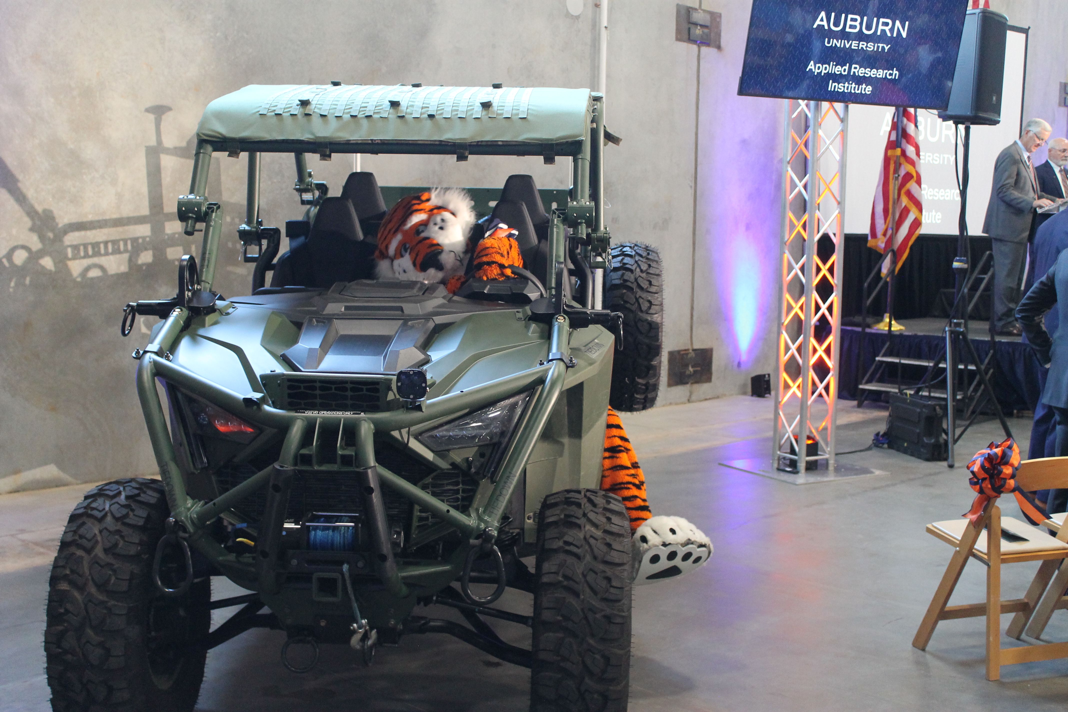 Green rugged off-road vehicle with a tiger plush in the driver's seat; to the right, a stage with an Auburn University banner, American flag, and two suited speakers.