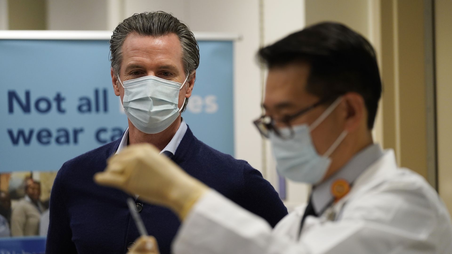 Photo of a masked Gavin Newsom watching a masked doctor with a needle