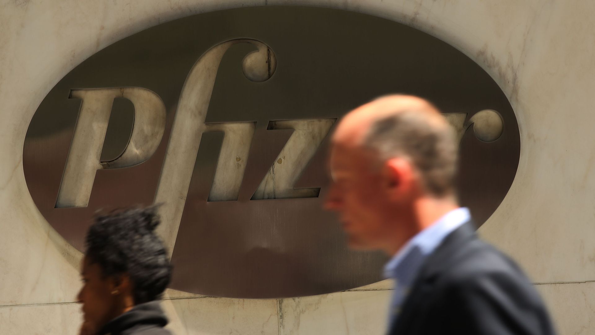 In this image, a man and a woman walk past a large metal logo for Pfizer