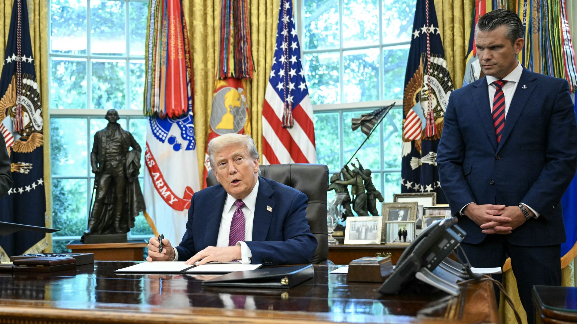 Sitting President Trump, wearing a navy jacket with a US flag pin, white shirt and purple tie, holds a black pen to sign executive orders next to a standing Defense Secretary Pete Hegseth (R), wearing a navy suit, white shirt and red-blue striped tie, in the Oval Office of the White House.