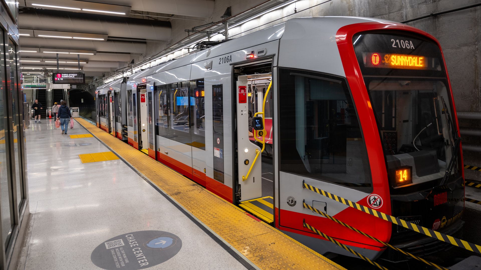 Modern light rail train with red and white colors at an underground station platform marked for Chase Center. Digital sign shows route to Sunnydale. Passengers walk along the platform.