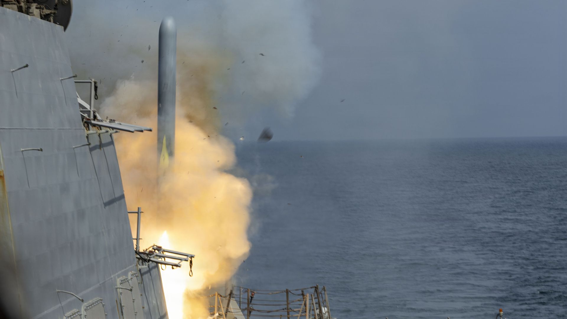 A side view of a gray warship at sea as a large orange flame and white smoke erupt from the deck and a missile zooms upward.