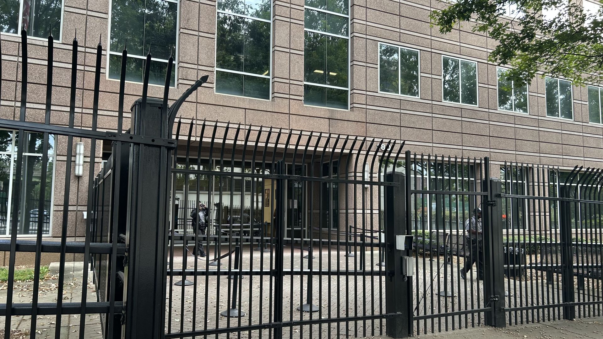 Black metal security fence with spikes in front of a brown stone office building with large windows and trees visible around it.