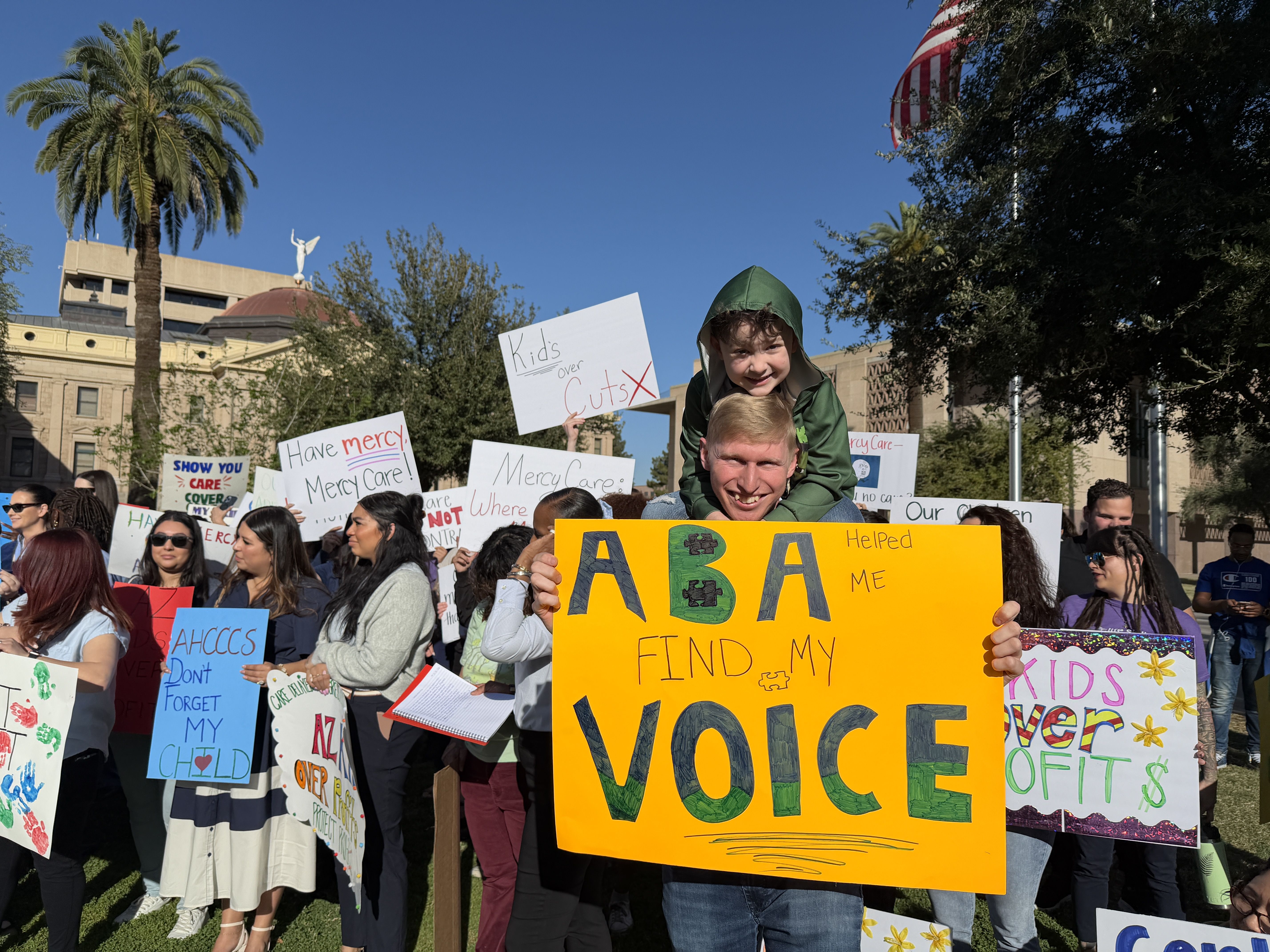 Man carrying child on shoulders holding bright yellow sign reading "ABA helped me find my voice" at outdoor protest with diverse group holding various signs under blue sky.