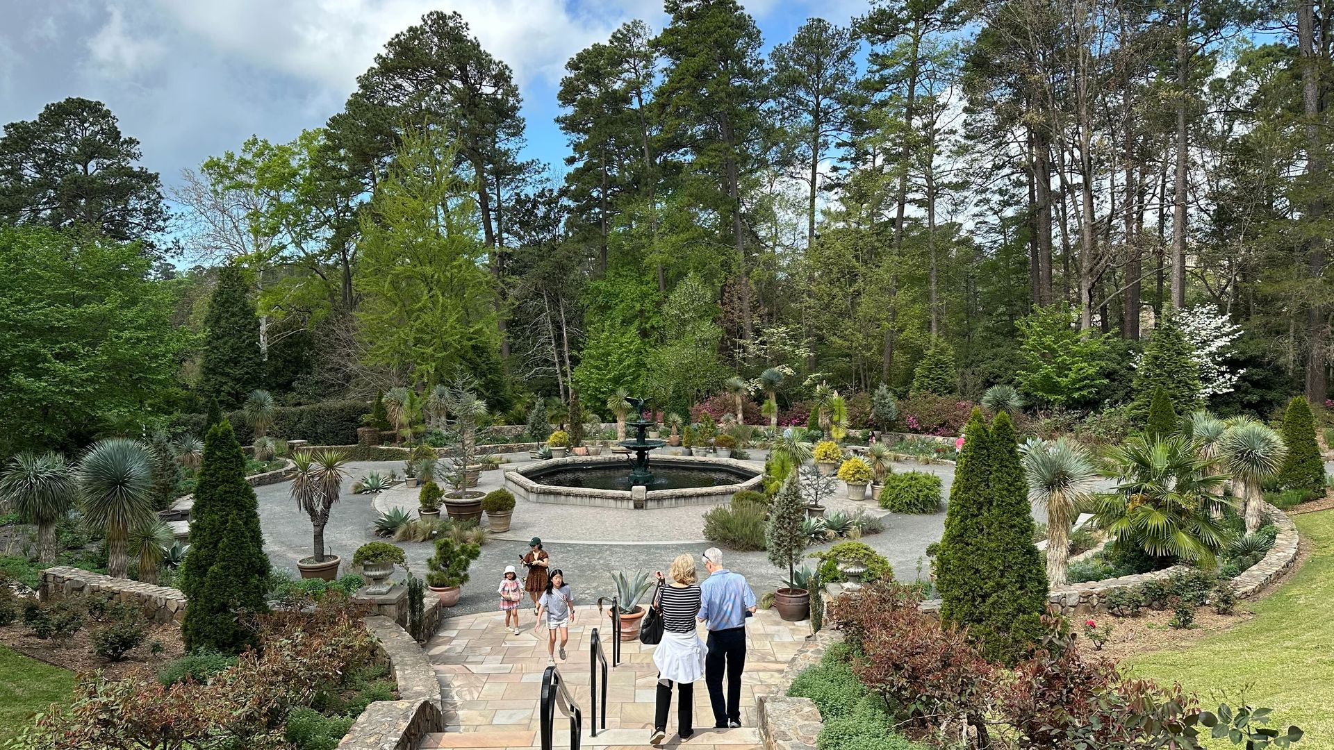 Formal garden with a circular fountain at center, surrounded by potted shrubs and topiary, stone steps and paths; visitors walk toward the fountain, tall trees and blue sky above.
