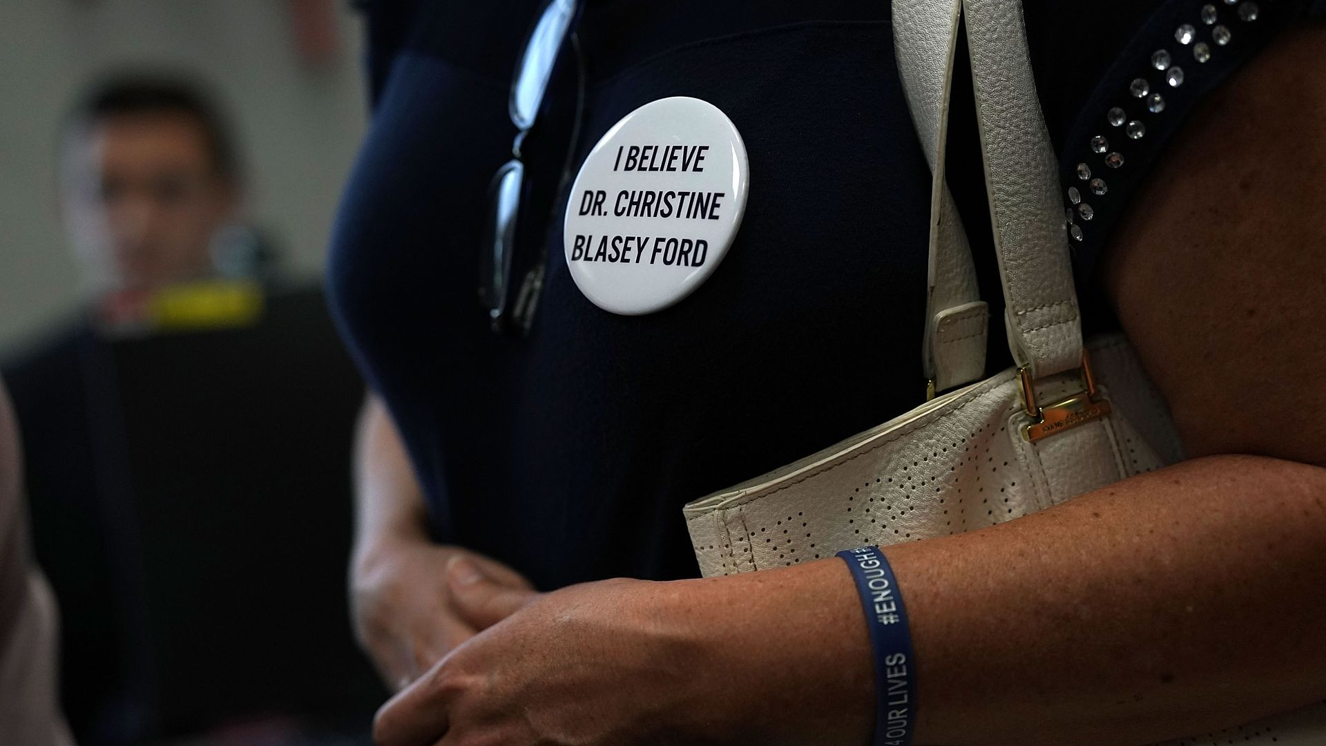 The chest of a woman wearing a dark blue shirt with a white round pin that says "I believe Dr. Christine Blasey Ford" 