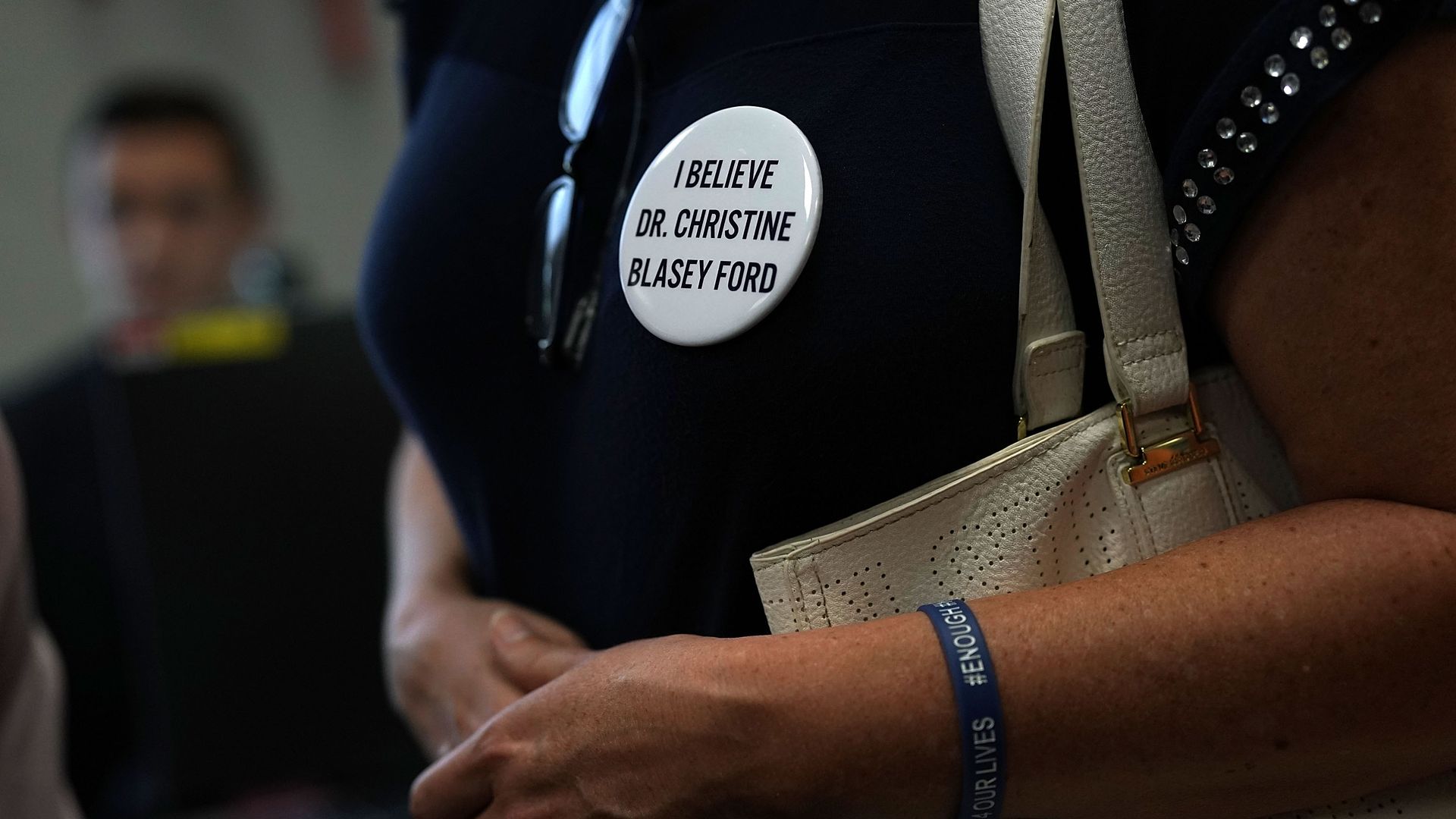 The chest of a woman wearing a dark blue shirt with a white round pin that says "I believe Dr. Christine Blasey Ford"