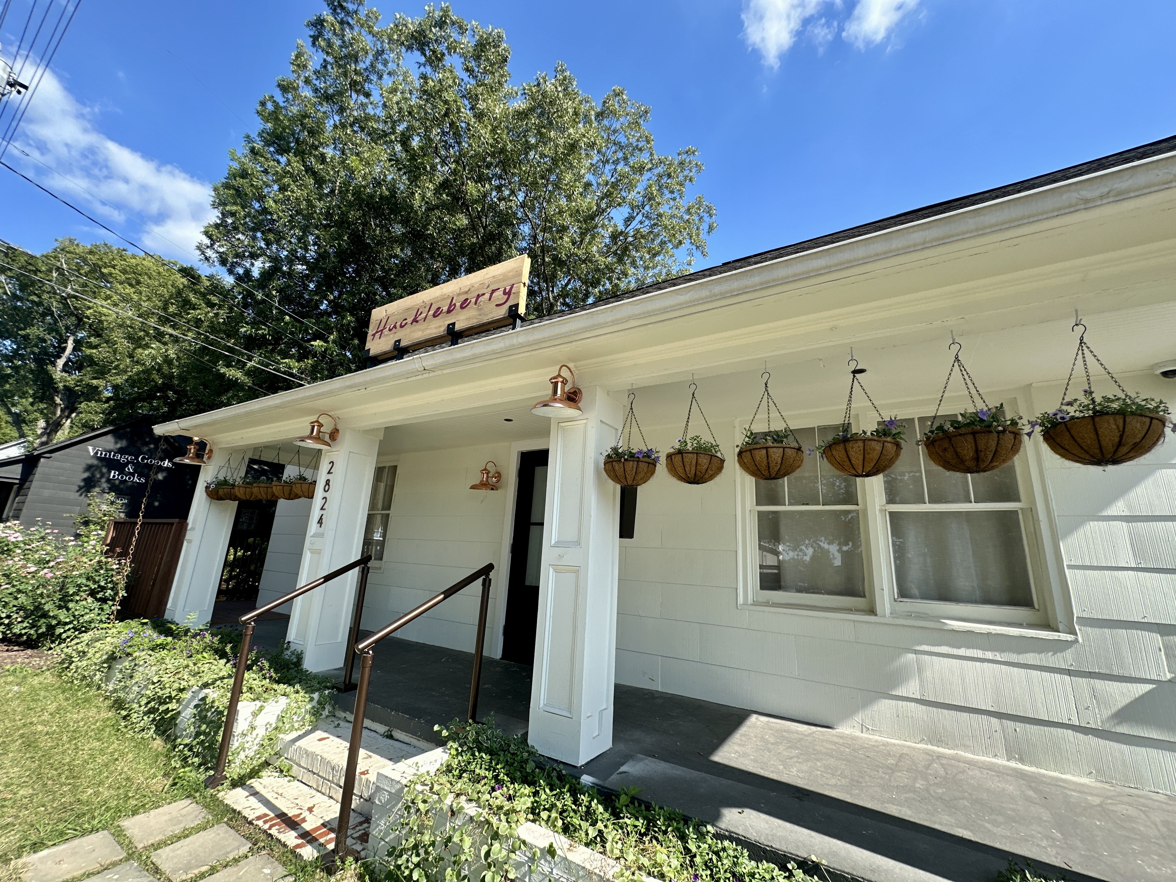 Sunny storefront with a white building and a wooden sign reading "Huckleberry" atop the roof. Hanging basket planters with purple blooms line the front; a large tree and blue sky behind.
