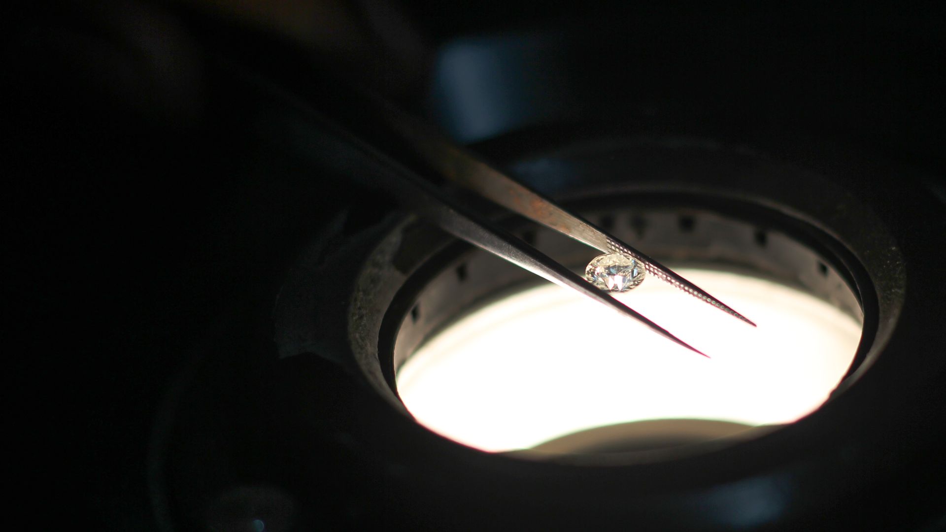 In this image, a small diamond is held between two long tweezers on top of a microscope.