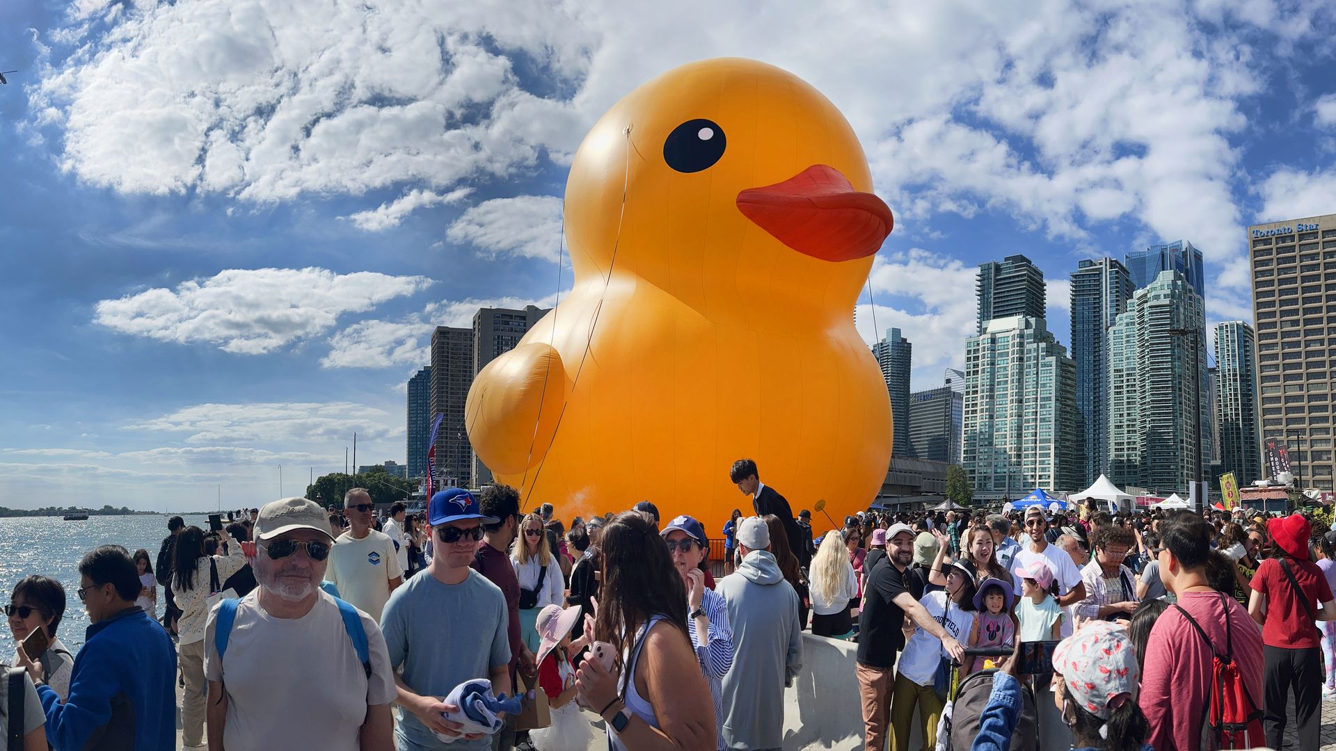 A giant inflatable yellow duck with buildings in the background and people in the foregroubd