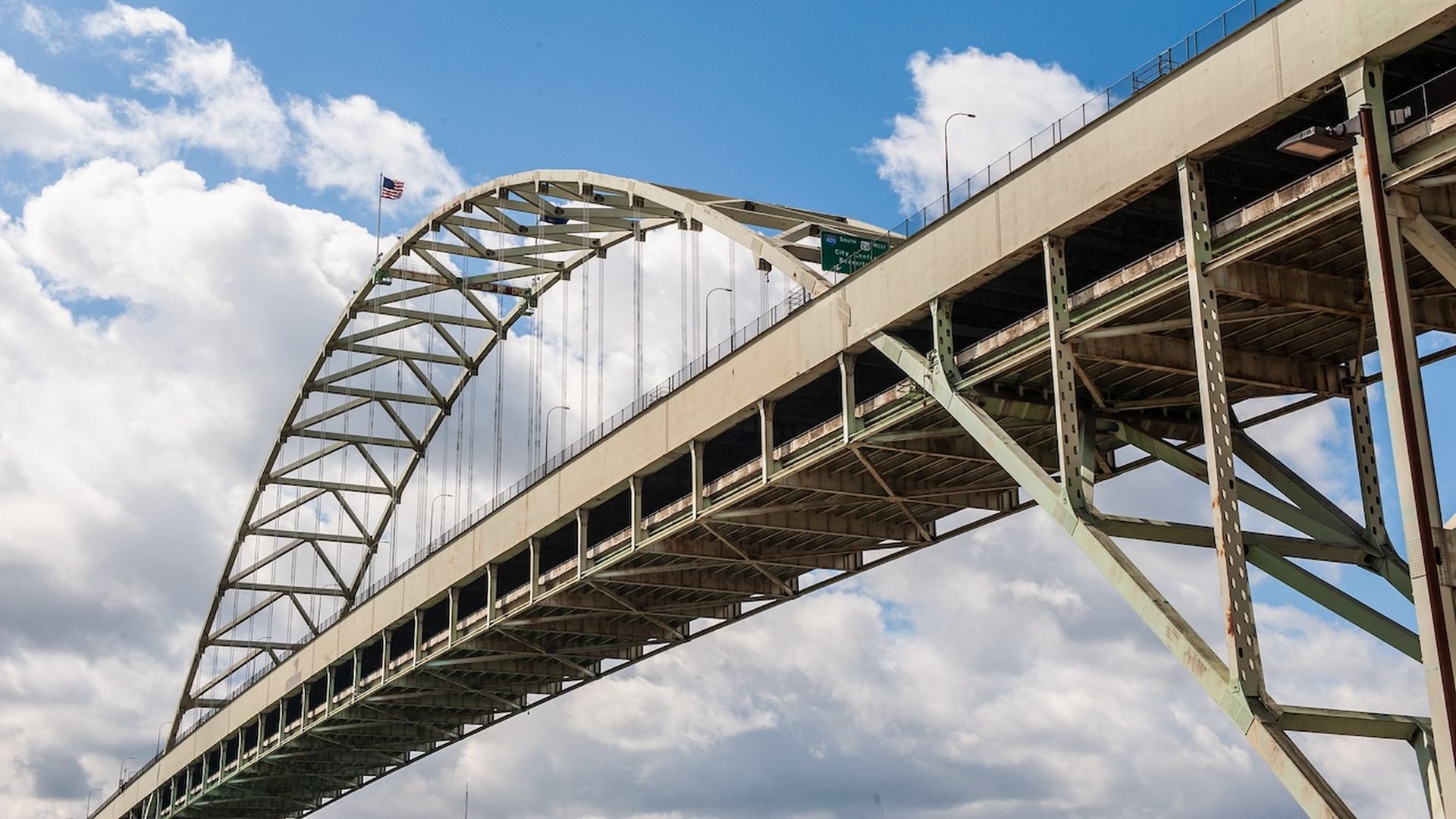 A large steel arch bridge with green-gray trusses and a roadway deck, seen from below. An American flag flies atop the arch against a blue sky with white clouds.