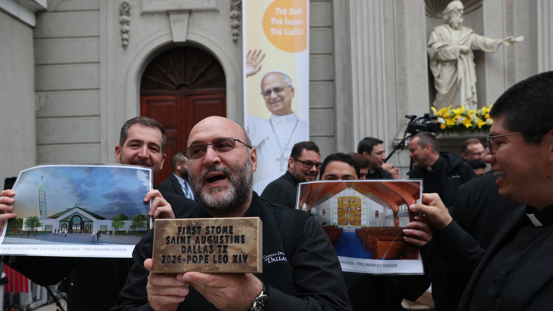 Men wearing Catholic clergy attire hold designs of Saint Augustine Church and a brick reading "First Stone Saint Augustine Dallas TX 2026 - Pope Leo XIV" outside a building with a banner featuring Pope Leo XIV in Istanbul, Turkey.
