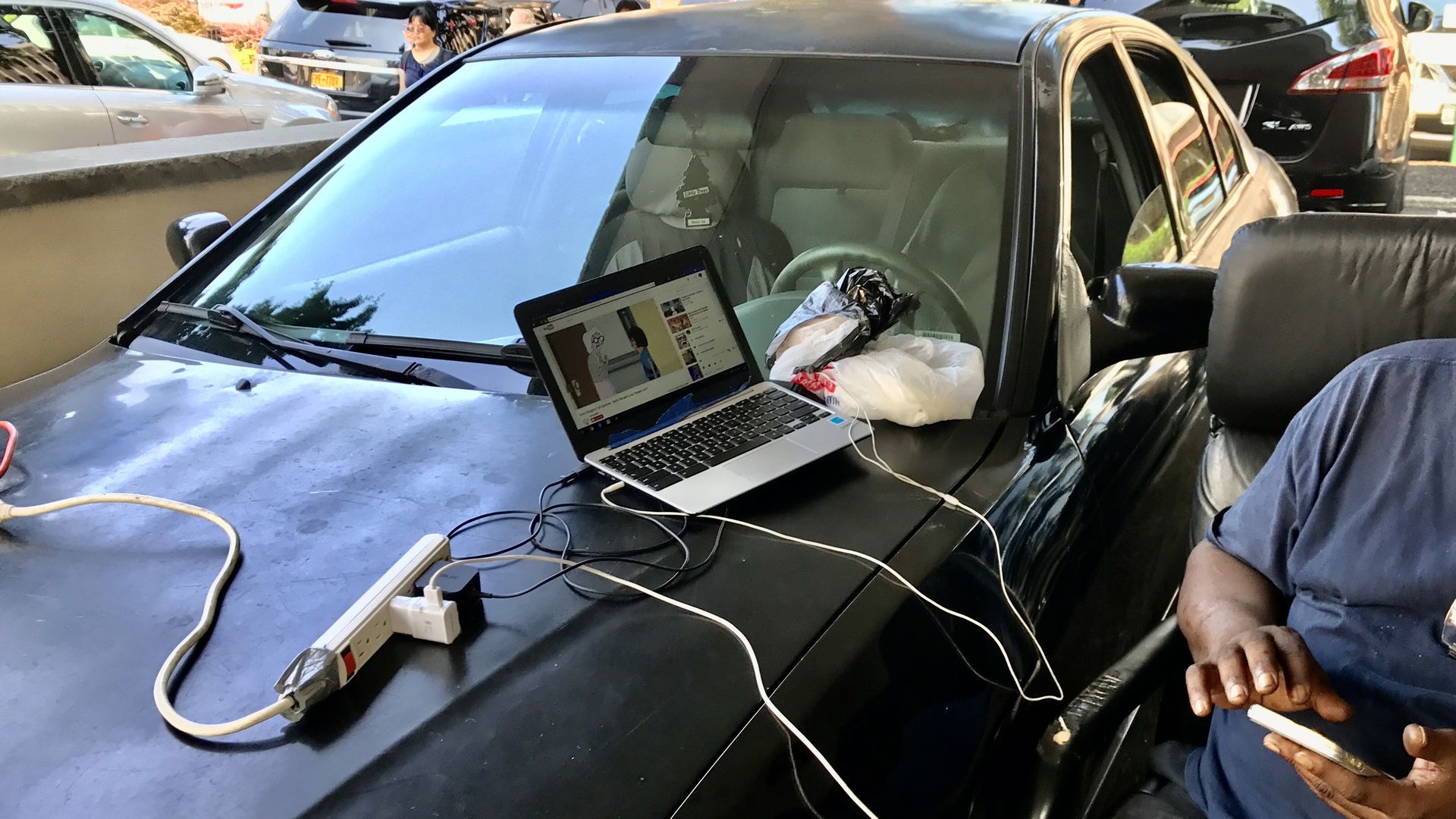 A man charges his laptop and phone while sitting next to his car and using the car's battery power for juice.