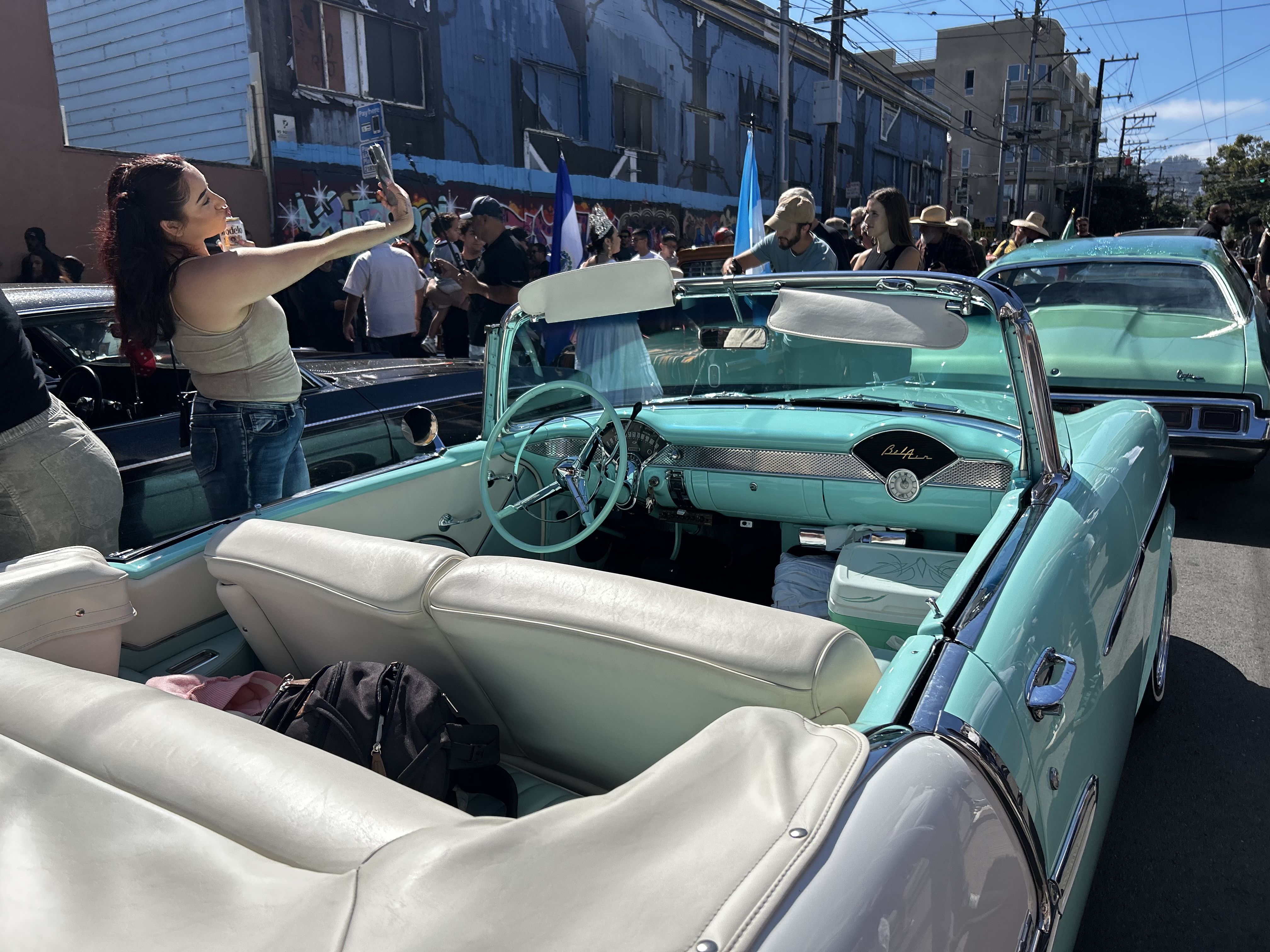Photo of two mint-colored lowriders parked on the street as a woman takes a selfie beside one