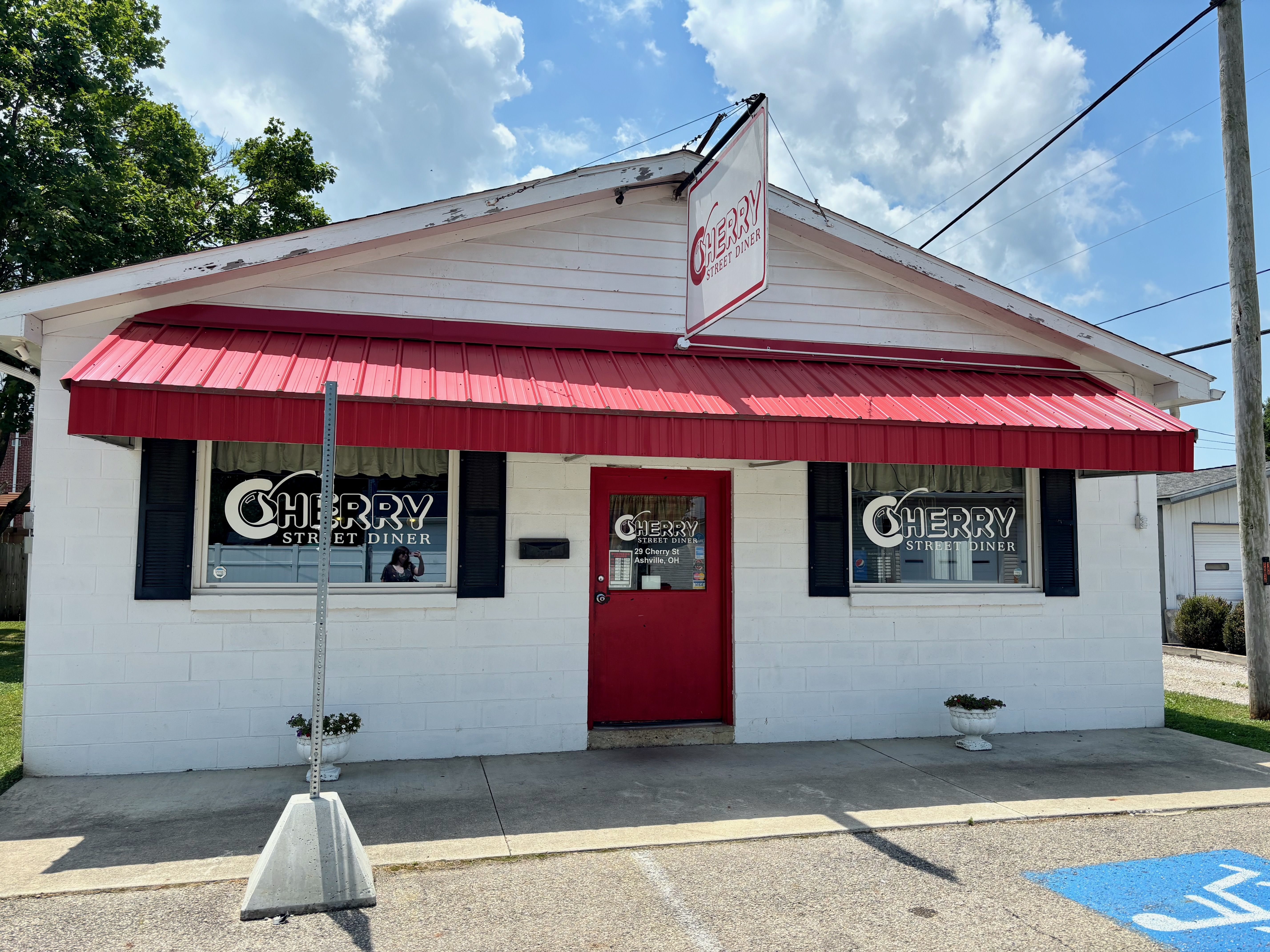 Front view of a small white diner with a red awning and red door. Windows display "Cherry Street Diner"; a hanging sign above reads it. Blue sky with clouds, trees, and power lines overhead.