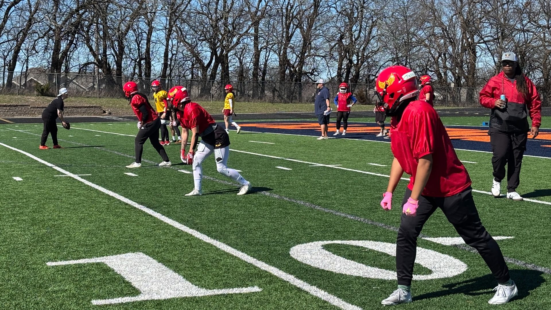 Young football players in red jerseys and helmets line up on a green turf field with white yard lines; a coach watches from the left as others in yellow practice jerseys stand in the background as coach Keke Blackmon walks among the formation in a gray Glory hat and red windbreaker. 