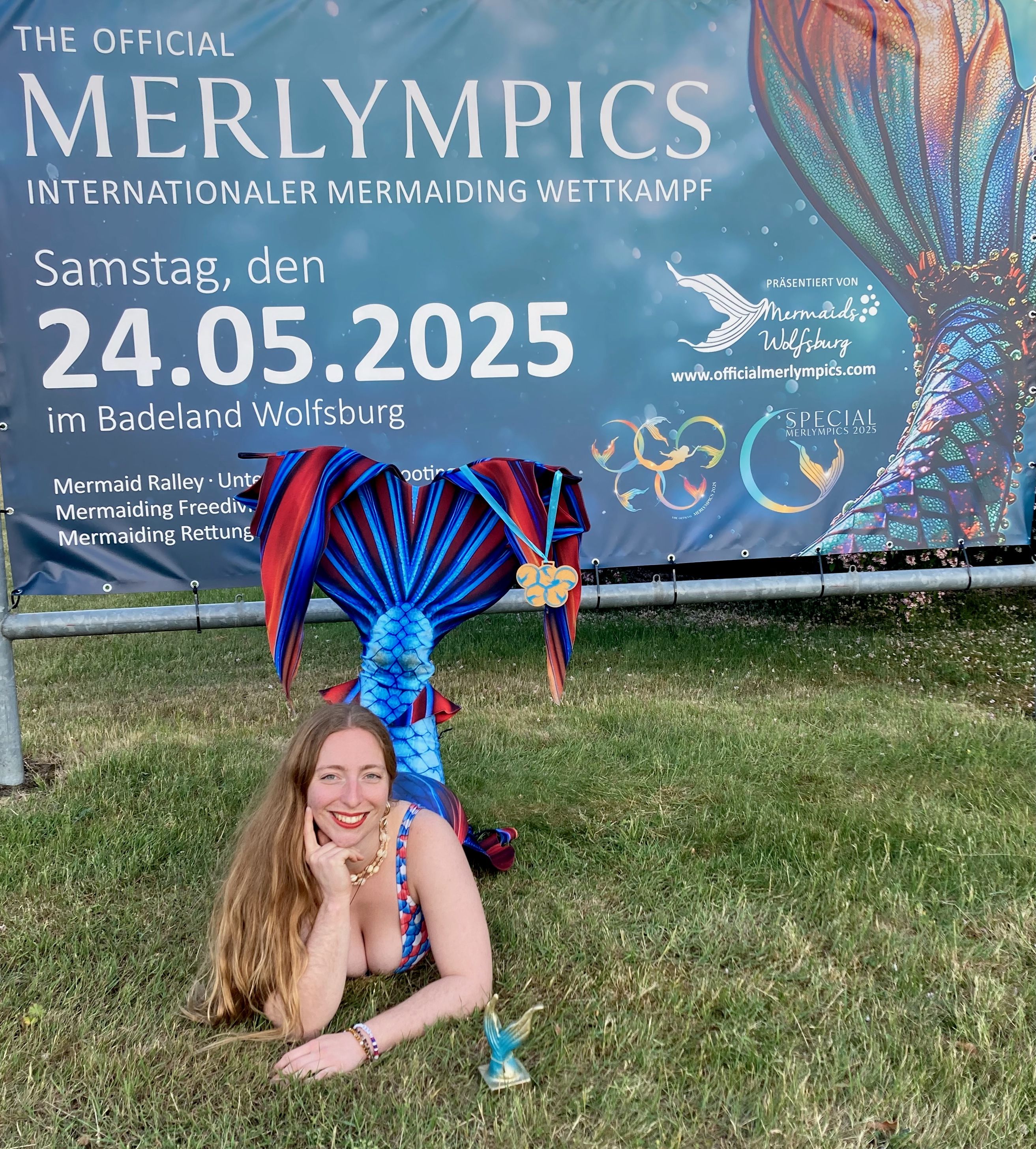 A woman dressed as a mermaid poses in front of a sign advertising the "Official Merlympics," with a gold medal hanging from her tail.