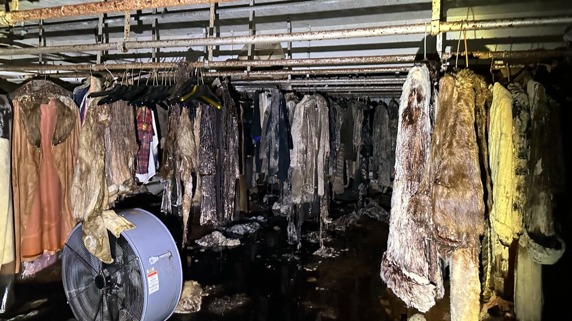 Dim, burned coat storage: rows of charred fur coats and jackets hanging from racks under a rusted ceiling; soot covers garments, with a large fan in the foreground.