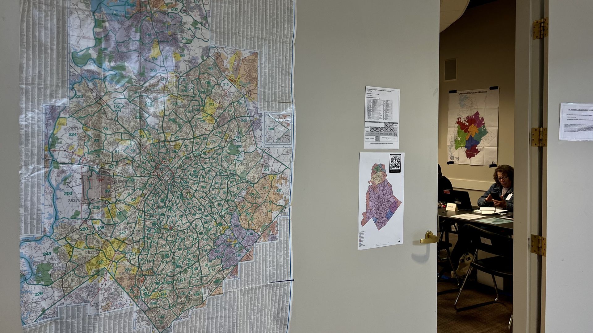 Large detailed map with numbered sections on a gray wall next to an open door showing a room with a person sitting at a desk with maps on walls, including a colorful district map.