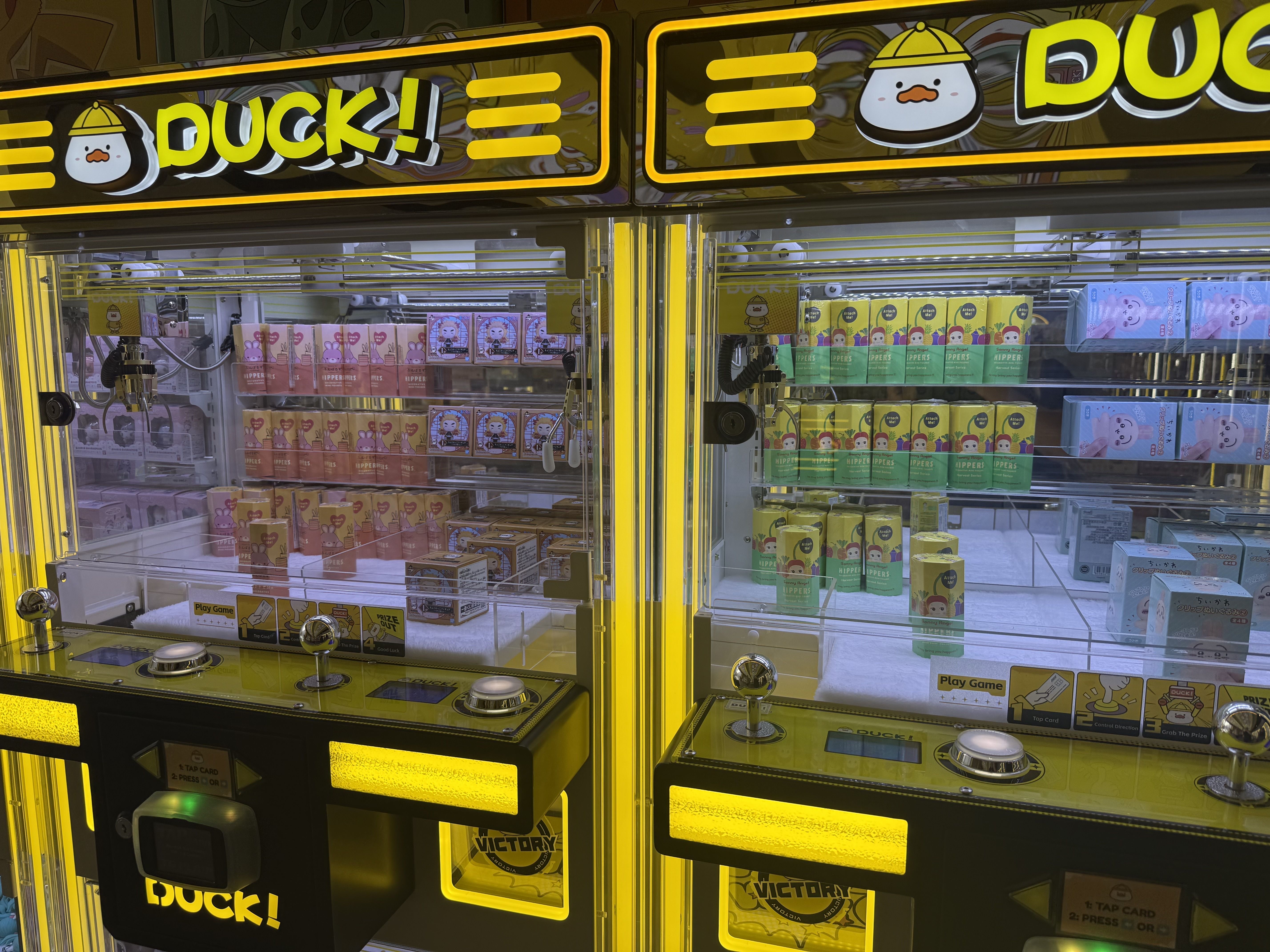 Bright yellow duck-themed crane game cabinet with neon borders, a large DUCK! sign and duck mascots, filled with pastel-colored prize boxes and packets behind glass.
