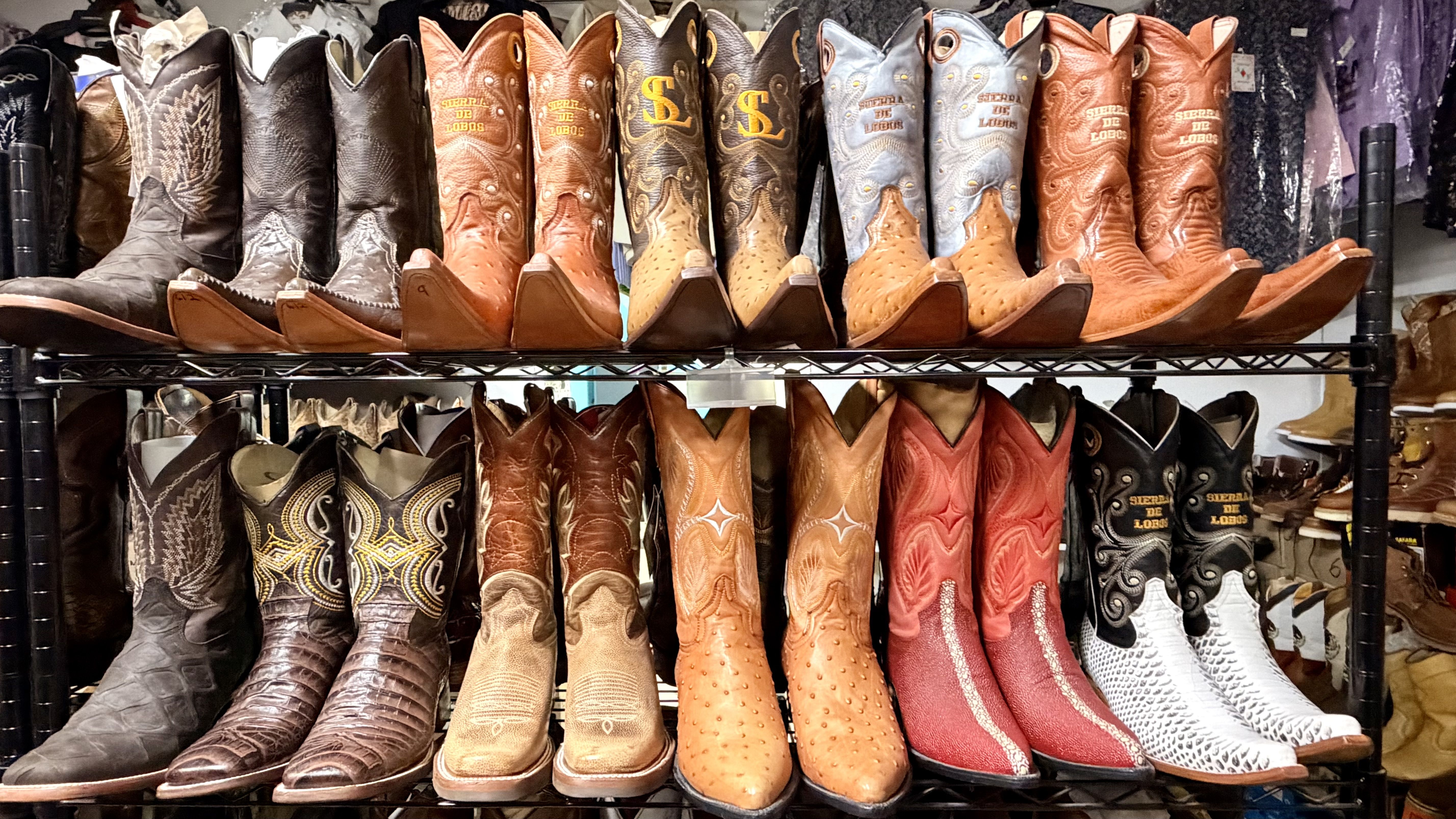Row of cowboy boots on metal shelves in a store, featuring brown, tan, black, and colored pairs with decorative stitching and pointed toes.