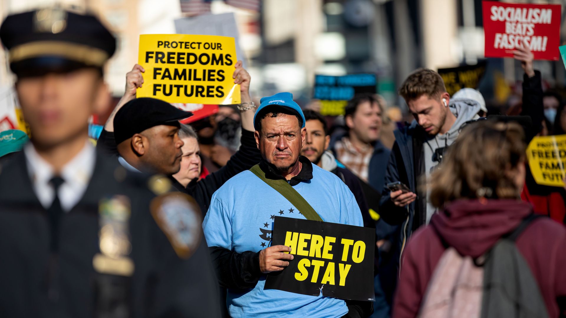 A man in a blue shirt holds a black sign with yellow letters that say "here to stay" during a protest in NYC. There are people all around him