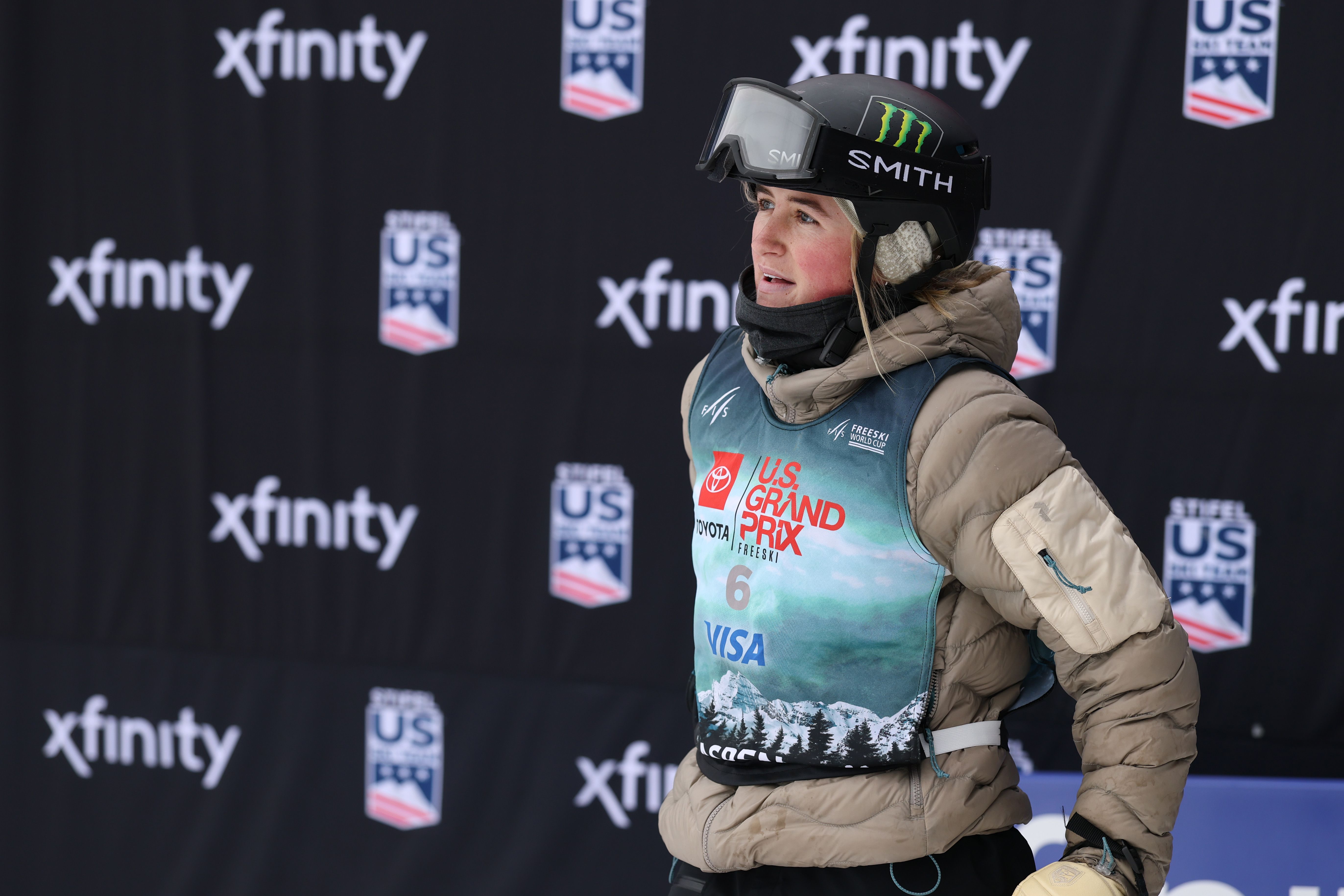 Female skier in tan jacket and black helmet with Monster and SMITH logos, wearing a blue bib with "U.S. Grand Prix" and sponsors, standing in front of black backdrop with "xfinity" and "US" logos.