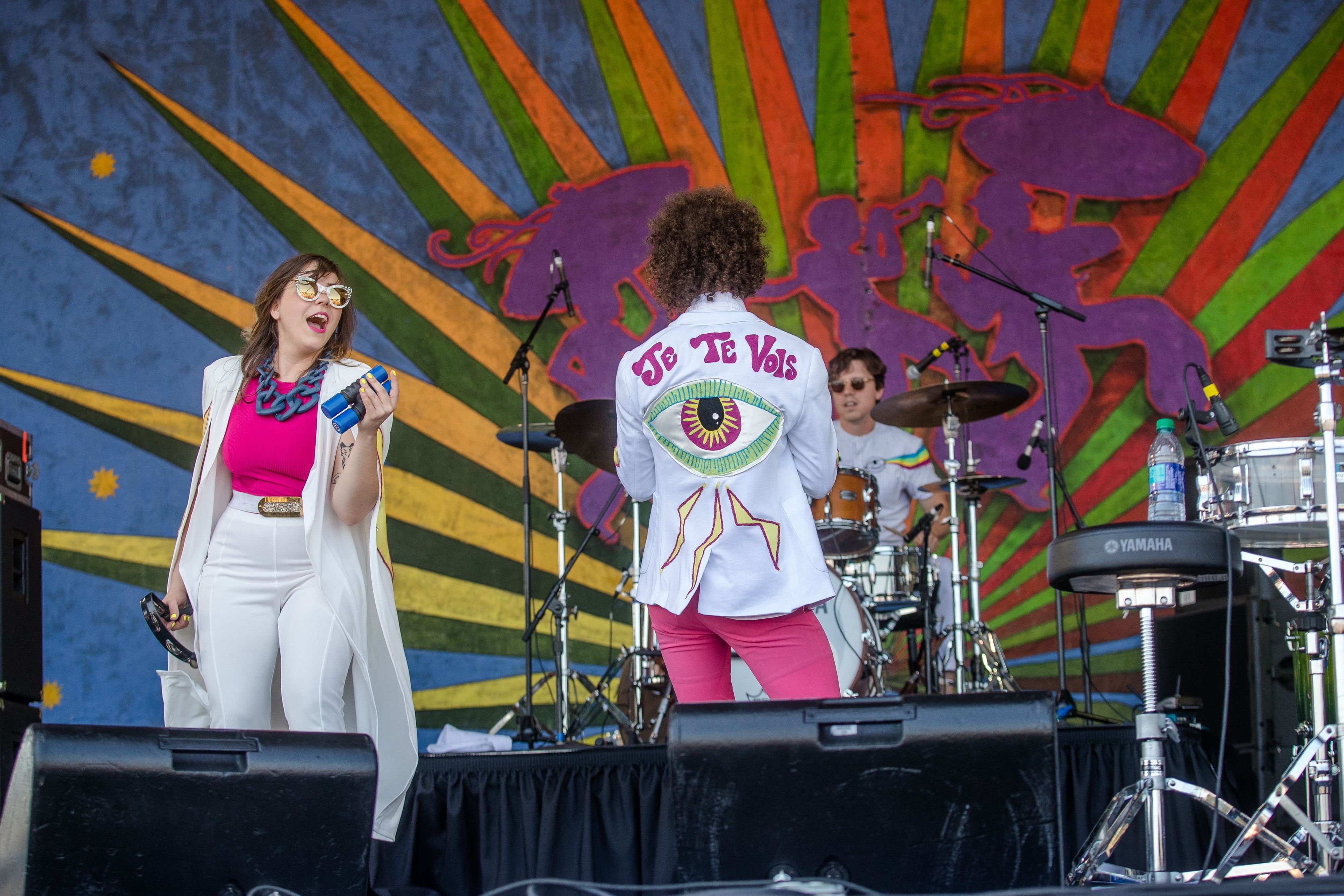 Sweet Crude performs on Jazz Fest's Gentilly Stage wearing customized white suits embroidered by Sigourney Morrison. Sam Craft stands at center stage with his back to the audience, showing an embroidered jacket that says "Je Te Vois" (or, "I see you") above a green and pink eyeball and lightning bolts.