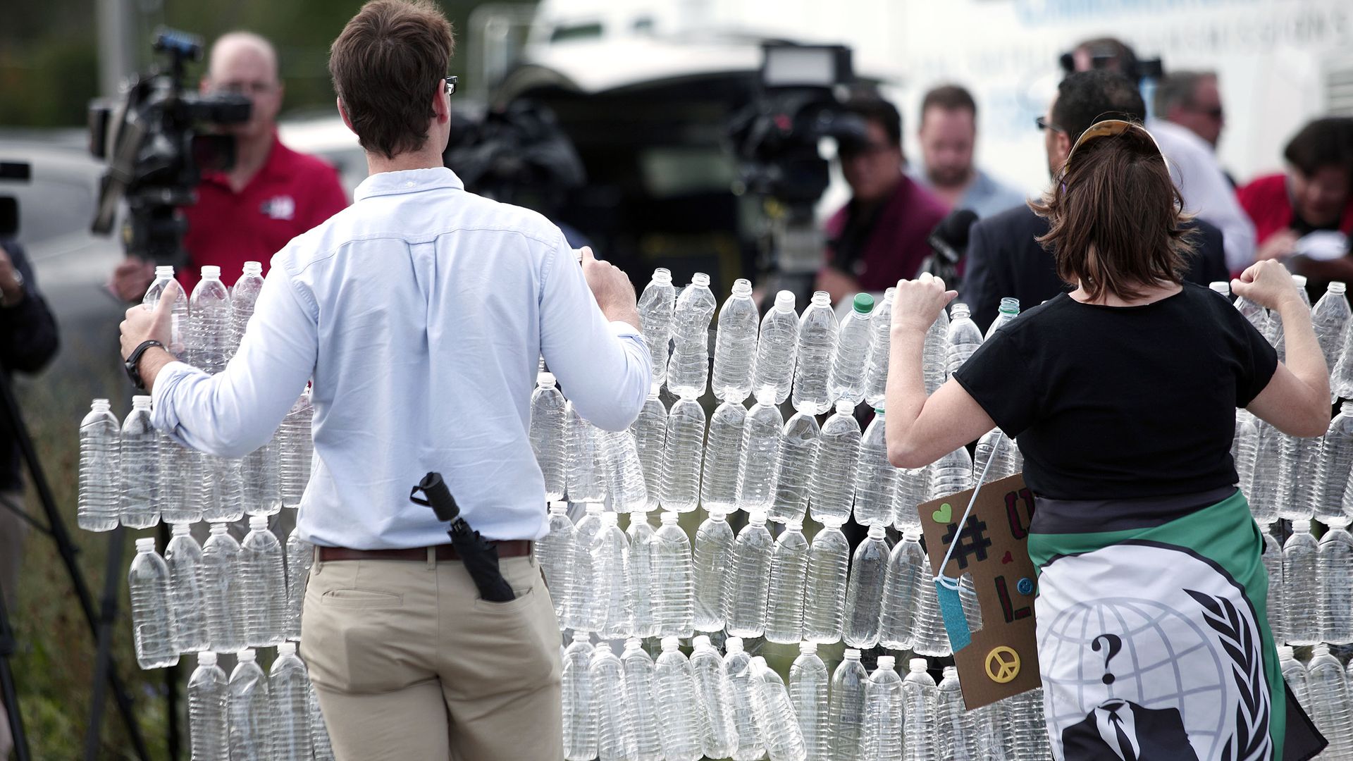 In this image, a man and a woman hold a connected chain of four rows of plastic water bottles while walking outside.