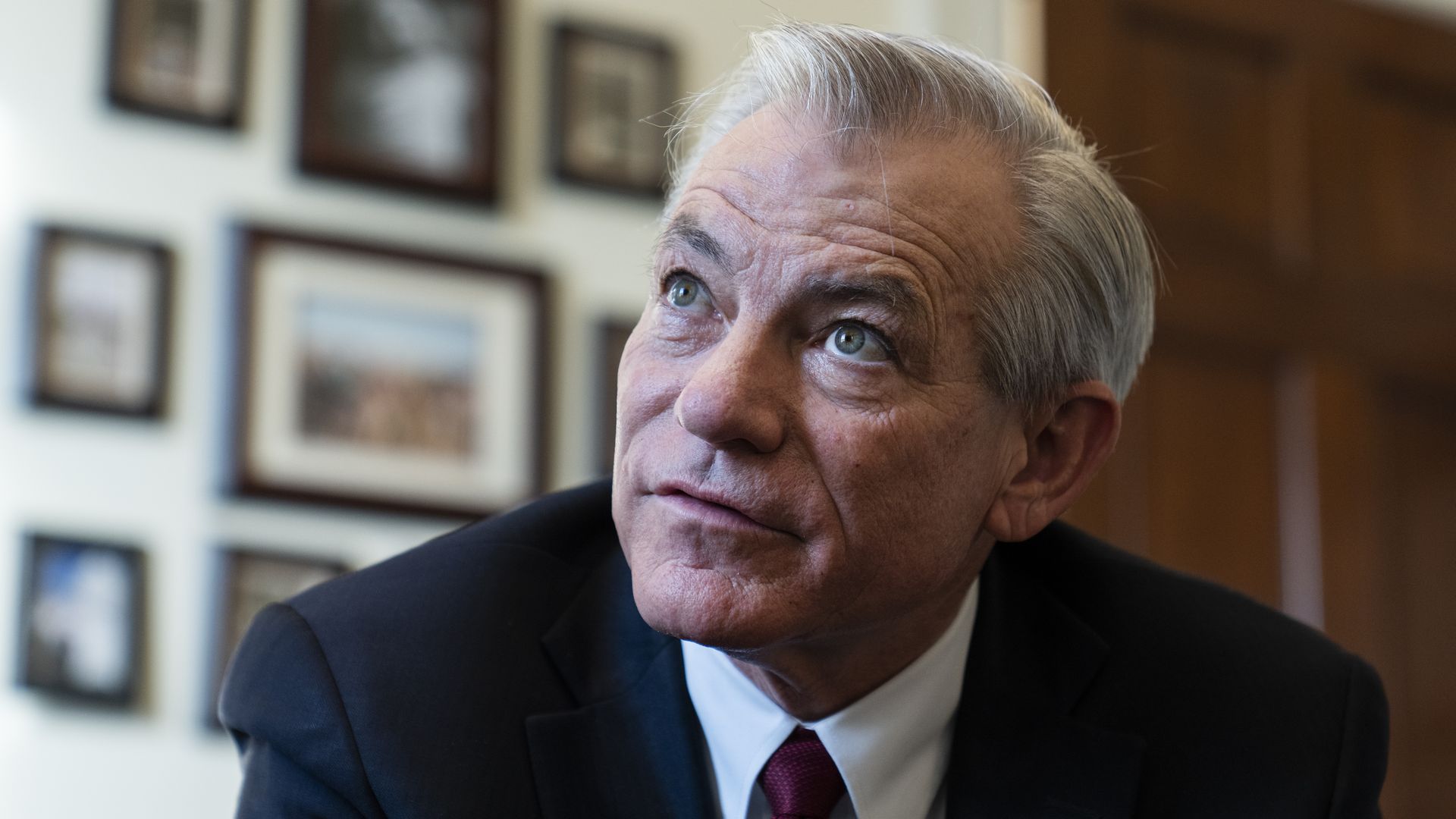 David Schweikert in a dark suit and red tie, looking thoughtfully upward. Blurred wall with framed photos in the background.