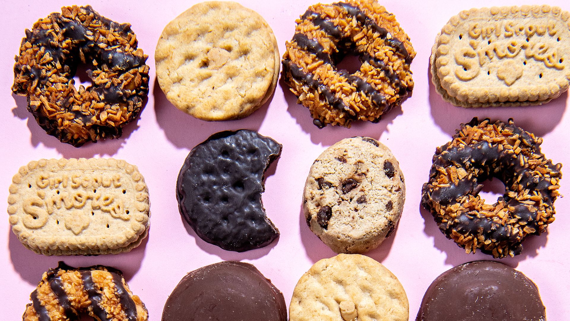 An array of Girl Scout cookies, including Smores, Samoas and Thin Mints, with one Thin Mint with a bite taken out of it.