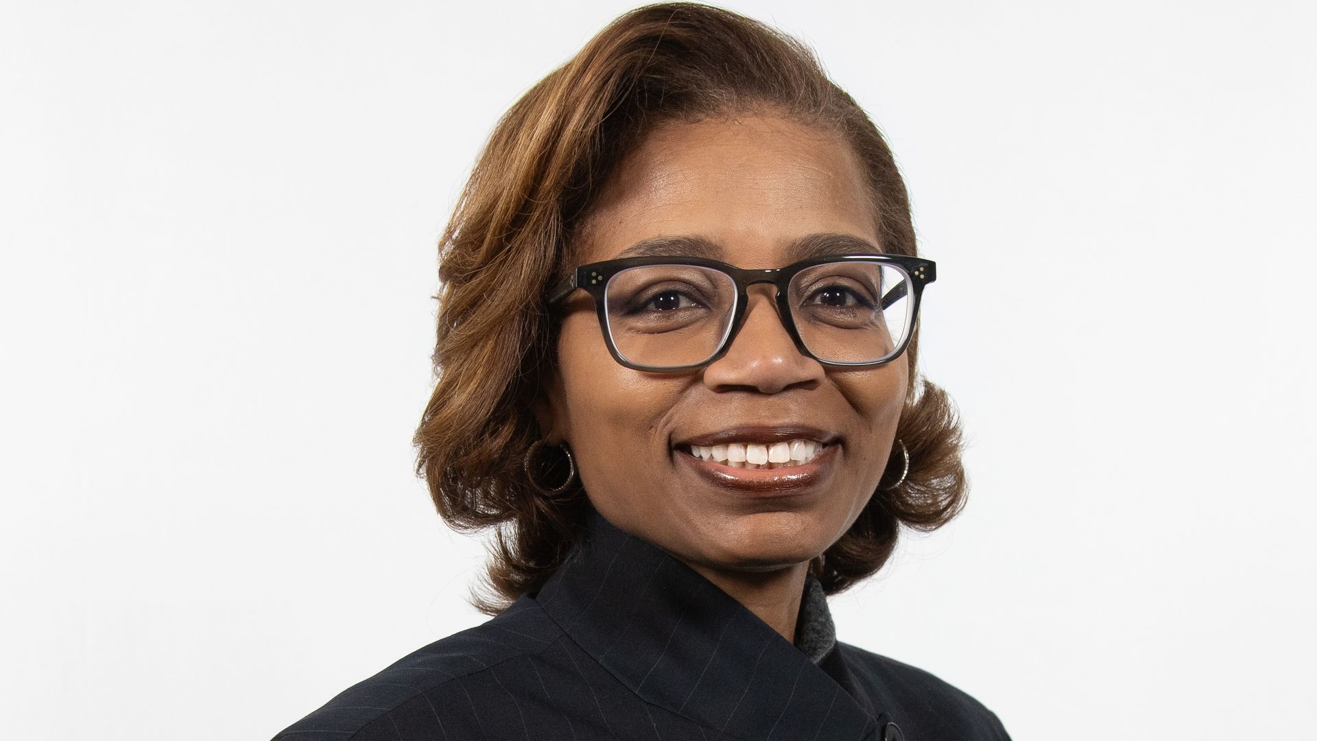 Portrait of a smiling Black woman with short brown hair and black glasses, wearing a dark pinstripe blazer, against a plain white background.