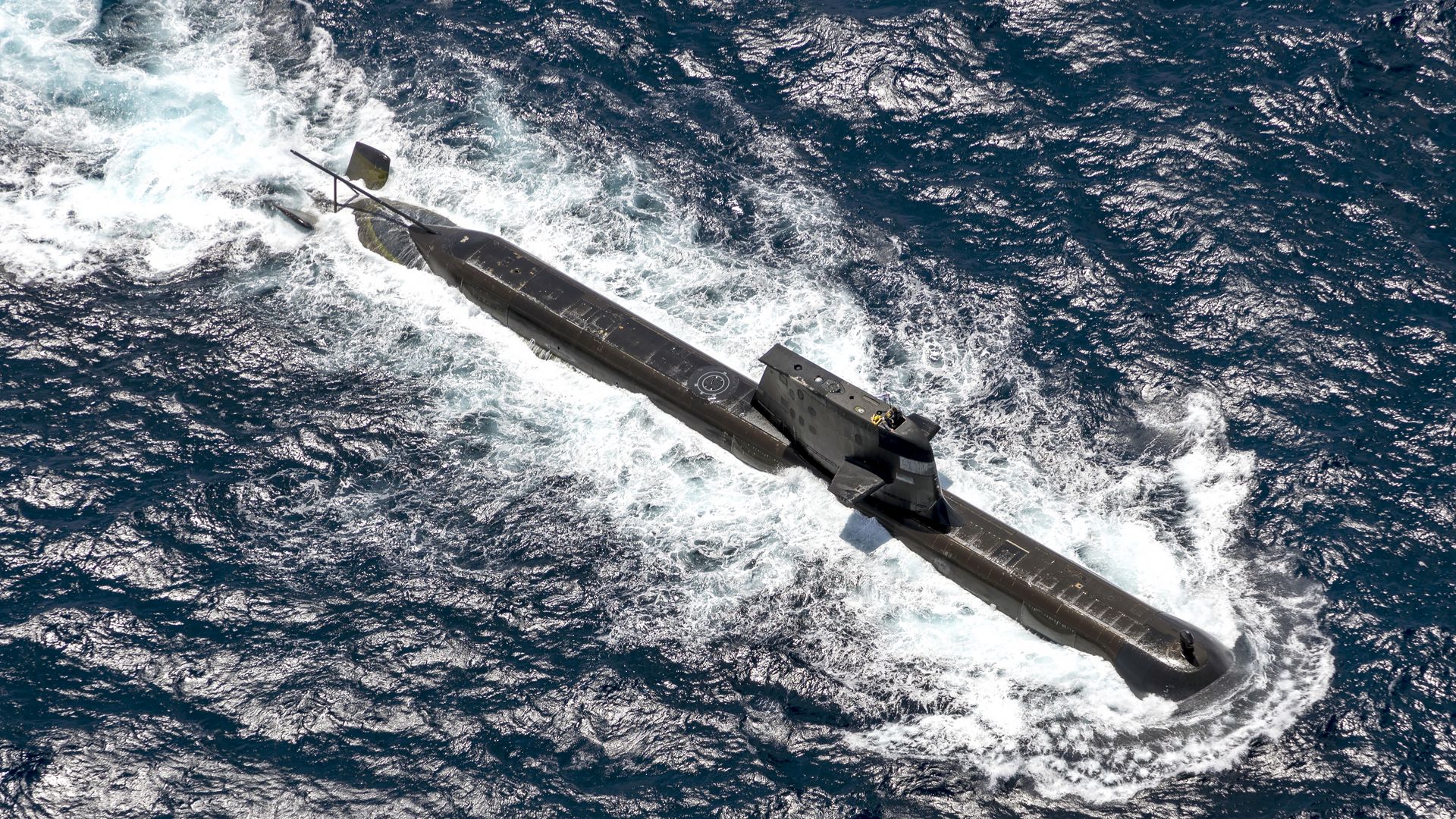 A black military submarine cruises at the ocean surface, creating white foamy wake against dark blue water under sunlight.