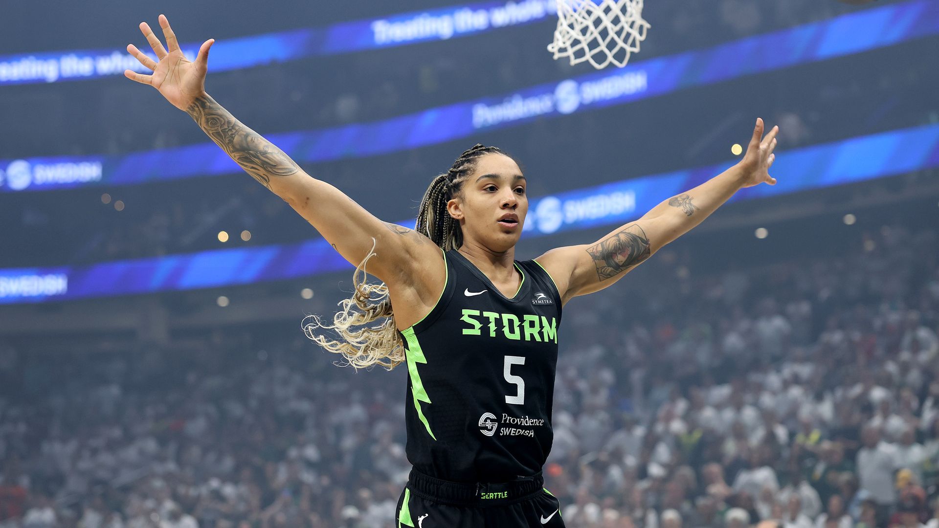 A basketball player holds out her hands wide as if to block passes or shots, in a crowded arena with a basketball net above her. She wears a jersey that says Storm and No. 5. 