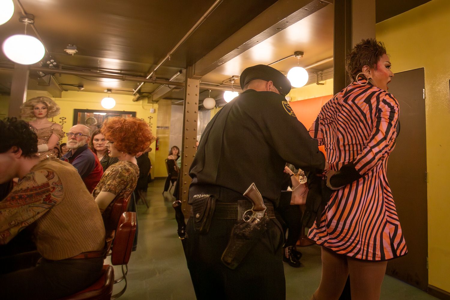 A drag queen in a pink and black zebra-striped coat stands near a uniformed officer in a yellow-lit backstage area, while seated audience members watch.